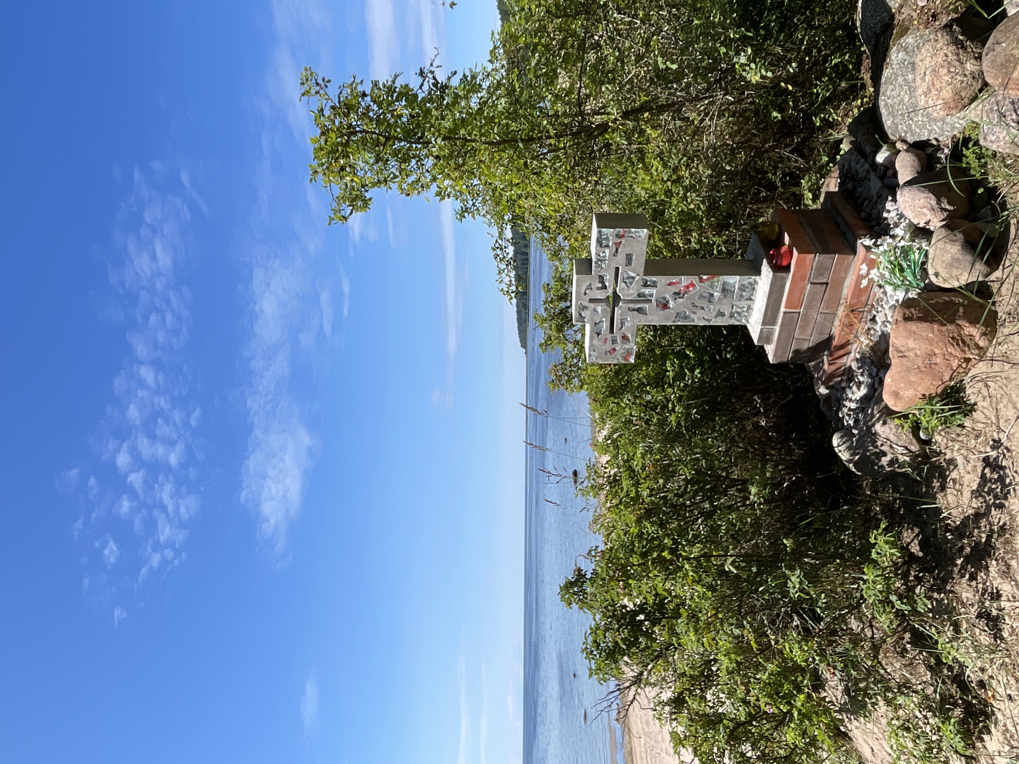 A cross made of concrete and mosaic tiles stands on a sandy shore next to a body of water. The cross is decorated with fragmented colorful glass and rests on a brick base. The surrounding area is covered in vegetation and rocks. In the background, the water stretches to the horizon under a bright blue sky with scattered white clouds. The scene appears to be a serene natural landscape, possibly a memorial or devotional site. The location is identified as Zelenaya Roshcha, Russia.
