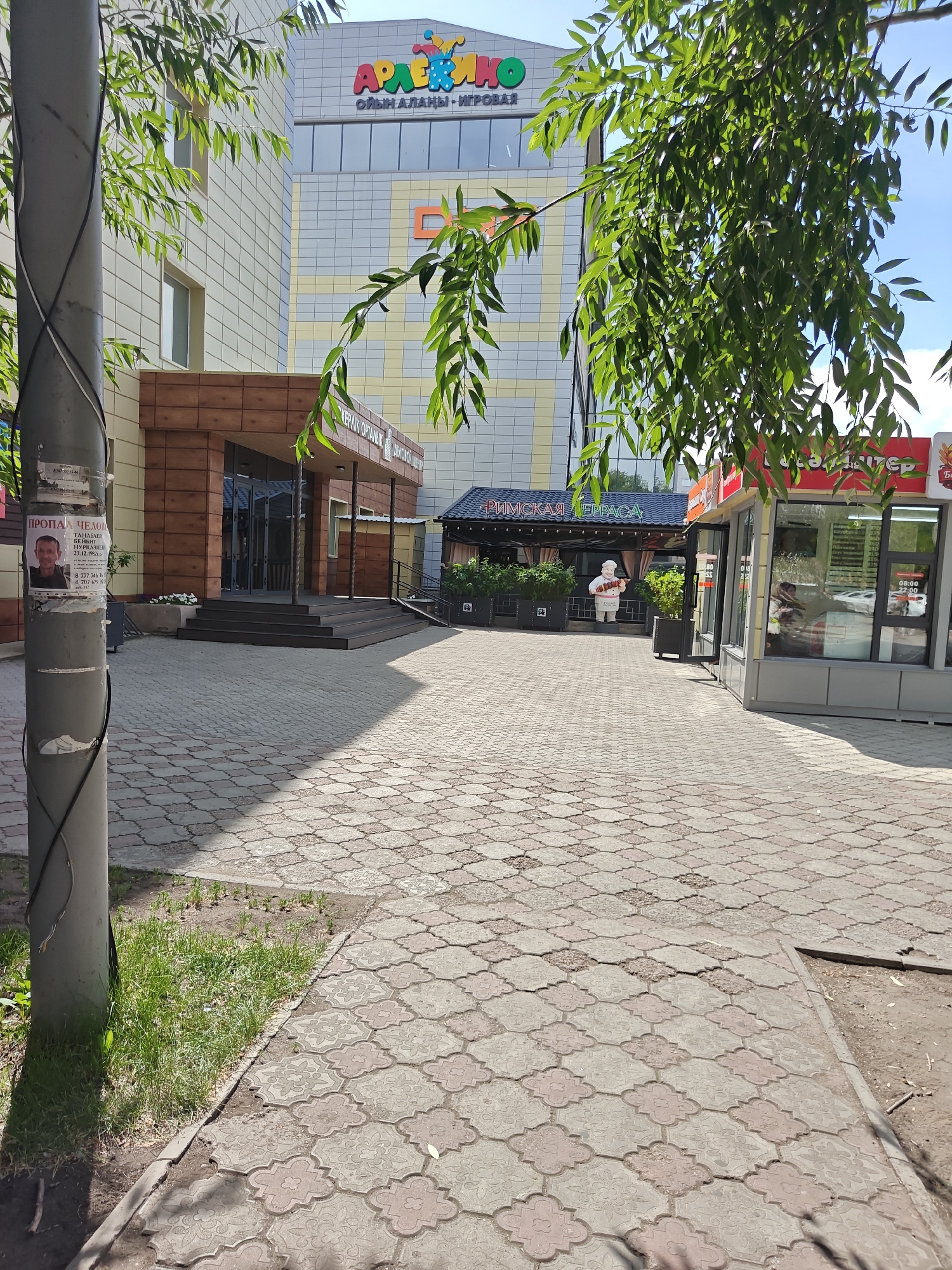 The image shows the exterior of a commercial building in Petropavl, Kazakhstan, specifically the "Arlekin" entertainment center. The building has a light-colored facade with orange and yellow accents and a prominent "Arlekin" logo at the top. In front of the building, there is a paved walkway made of interlocking concrete pavers with a decorative pattern. To the left of the walkway, there is a lamppost with a poster attached to it, displaying a missing person notice. A tree with green leaves partially obstructs the view of the building. To the right of the entrance, there is a small kiosk or shop with a red awning and large glass windows. Further to the right, there is another covered structure with a dark blue roof and a sign that reads "РИМСКАЯ ТЕРРАСА" (Roman Terrace). A statue of a chef is visible outside this structure, holding a pizza. The sky is blue with some scattered clouds. The overall scene depicts an outdoor area of a public place, likely during daytime.
