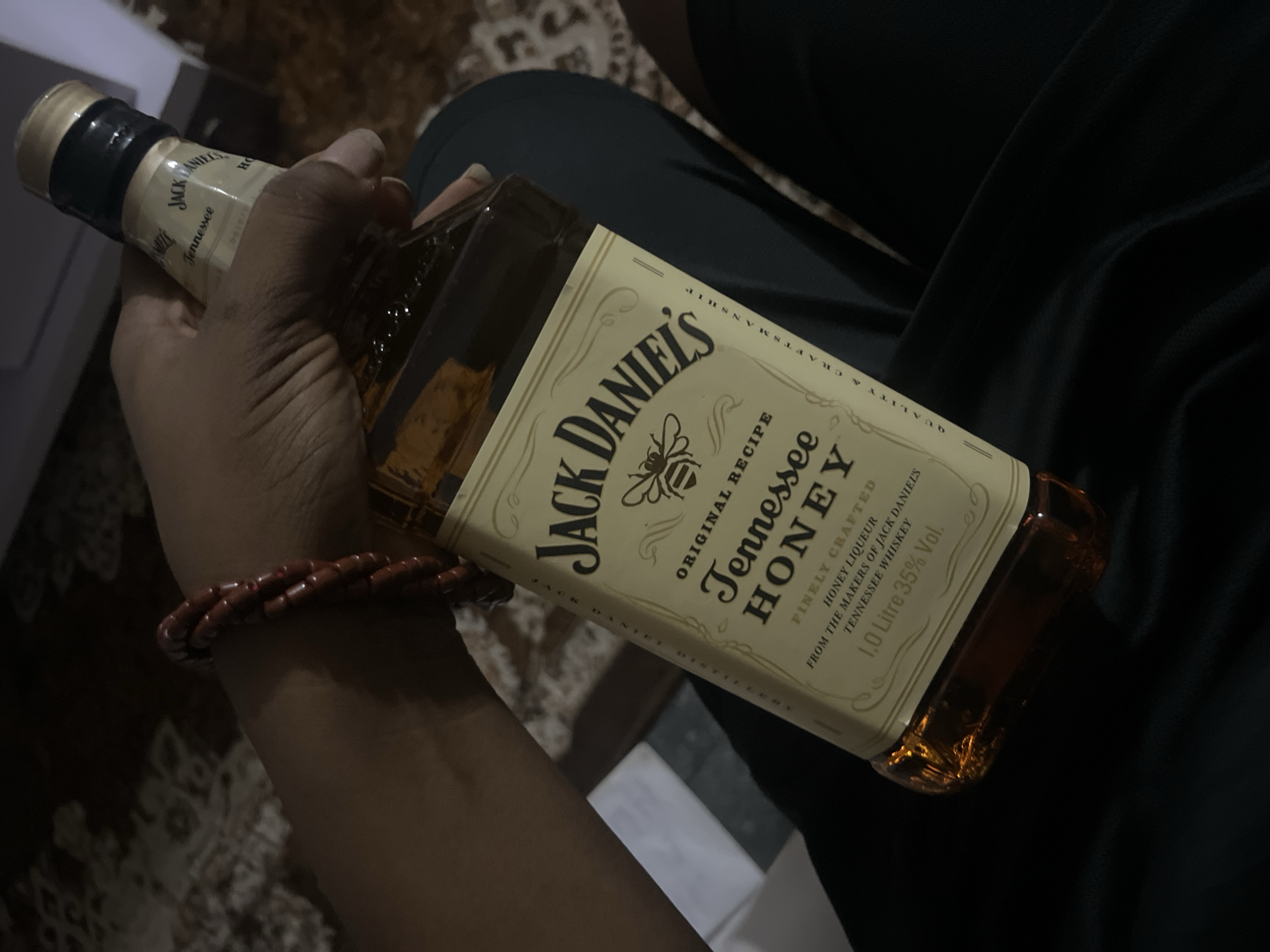 A bottle of Jack Daniel's Tennessee Honey whiskey is being held in someone's hand. The bottle is labeled with "Jack Daniel's", "Original Recipe", "Tennessee Honey", and "1.0 Litre 35% Vol". The person holding the bottle is wearing a beaded bracelet. The background includes a dark garment. The location is unknown.