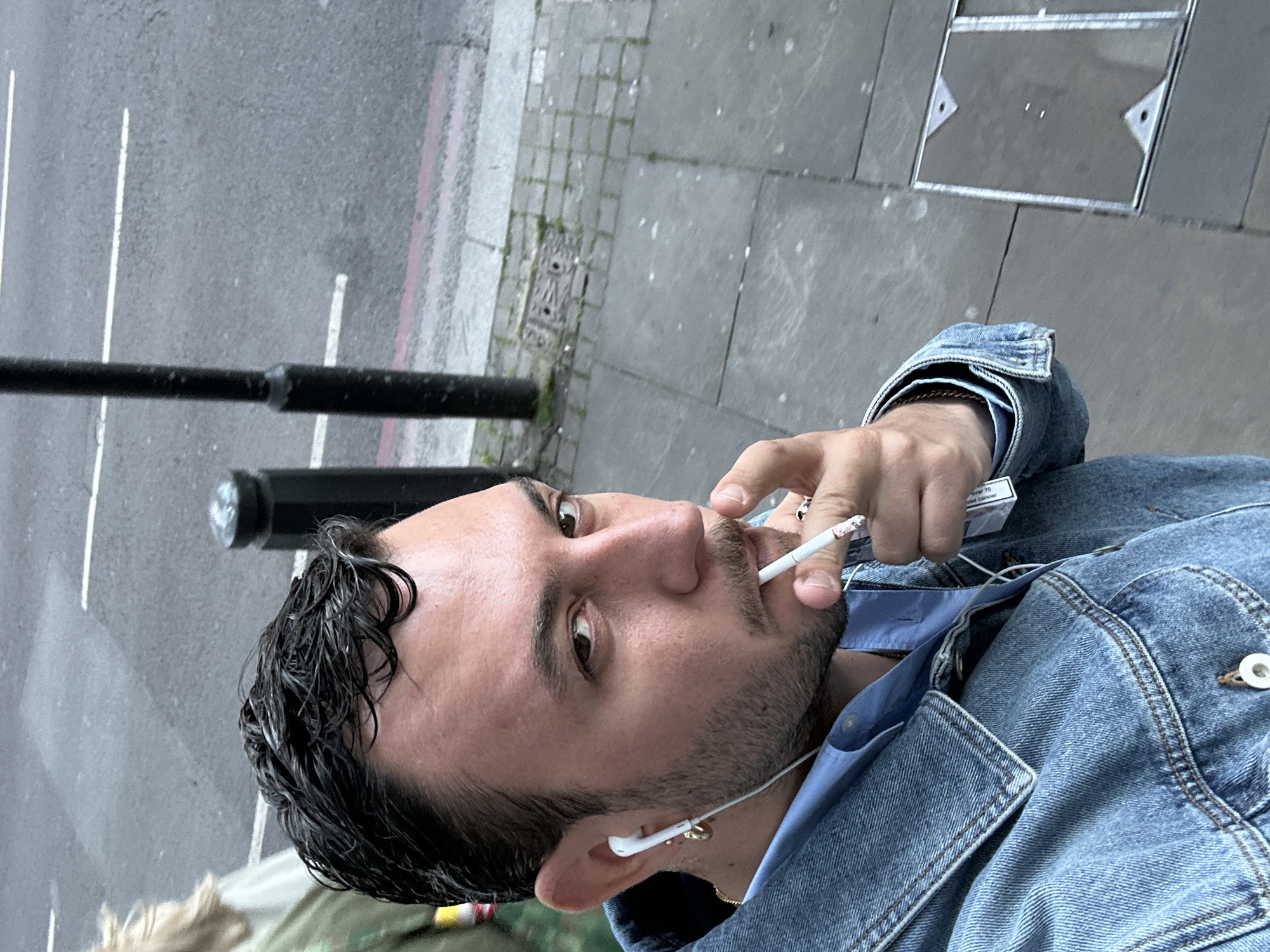 The image captures a close-up, rotated view of a man on an urban street, likely in London, United Kingdom.
The primary subject is a man in his mid-20s to early 30s, positioned in the lower-left to central frame. He has dark, wavy hair that appears wet, a short beard, and a mustache. He is wearing a denim jacket over a light blue collared shirt. A white wired earbud is visible in his right ear, along with a small gold hoop earring. He is actively smoking a lit cigarette, holding it between his fingers near his mouth. In his hand, he also holds a white cigarette pack.
The background shows a street and a sidewalk from an elevated perspective. The street features asphalt pavement with white painted lines and a thin red line along the curb, indicative of traffic regulations. The sidewalk consists of grey square paving stones, some with visible weeds growing in cracks, and a dark metal manhole cover with triangular accents. A black lamppost or pole is partially visible on the left side of the frame.
The lighting suggests it is daytime, possibly overcast, as there are no harsh shadows. The man's gaze is directed upwards and to the side, implying he is looking at something off-camera. His expression is neutral.
Visible text on the cigarette pack includes a health warning, with phrases like "SMOKING KILLS" and "tobacco smoke contains over 70 cancer-causing substances," along with the number "20" and "Cigarettes."