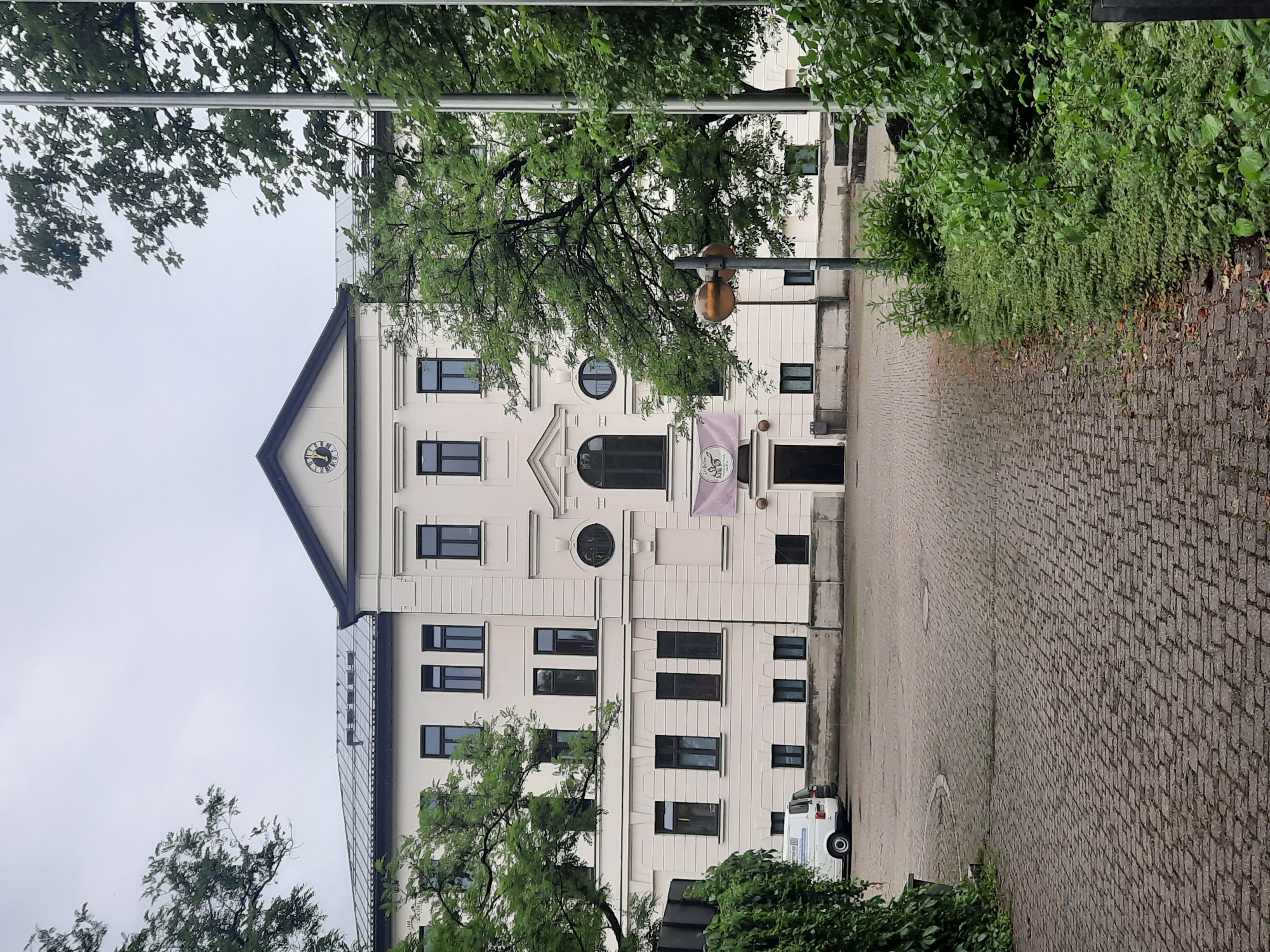 The image depicts the exterior of a cream-colored, multi-story building with a dark roof and a triangular pediment. A clock face with Roman numerals is visible on the pediment. The building has numerous dark-framed windows. A light pink banner with text and an illustration hangs above a dark entrance. To the left of the building, a white van is parked on a cobblestone driveway. The area is surrounded by lush green trees and foliage. The scene suggests an overcast day. Based on the style of architecture and vegetation, the location is likely a public building or an institution in Rosenheim, Germany.