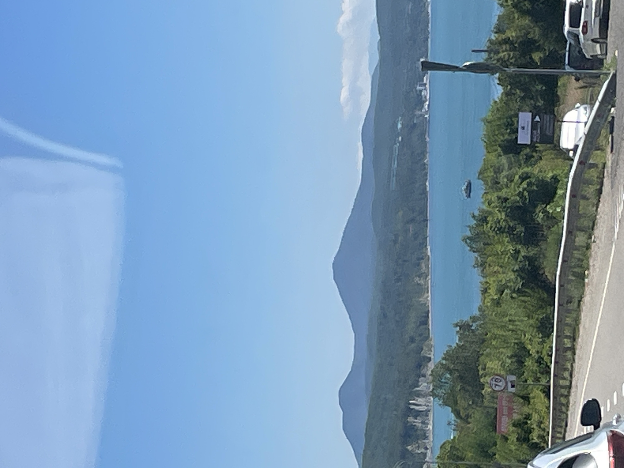 The image captures a scenic coastal view with mountains in the background. A bright blue body of water, likely the Black Sea, dominates the middle ground. Along the coastline, a dense green forest covers rolling hills that transition into rugged cliffs. In the distance, two distinct mountain peaks rise against the clear blue sky, with a faint cloud formation streaking across the upper right portion of the frame.
In the foreground, a paved road curves to the left, bordered by a metal guardrail. A silver car is partially visible on the left side of the road, suggesting the perspective is from another vehicle. To the right of the road, a group of trees and foliage are visible, with a speed limit sign showing "70" and another sign with an arrow pointing to the right. Further down the road, two white cars are parked or stopped.
A small boat is visible on the water, and in the far distance, structures that appear to be buildings are scattered along the coastline. The overall impression is a sunny day with good visibility, offering a panoramic view of the natural landscape. The scene likely depicts a section of the coastal road near Dzhubga, Russia, known for its beautiful mountain and sea vistas.