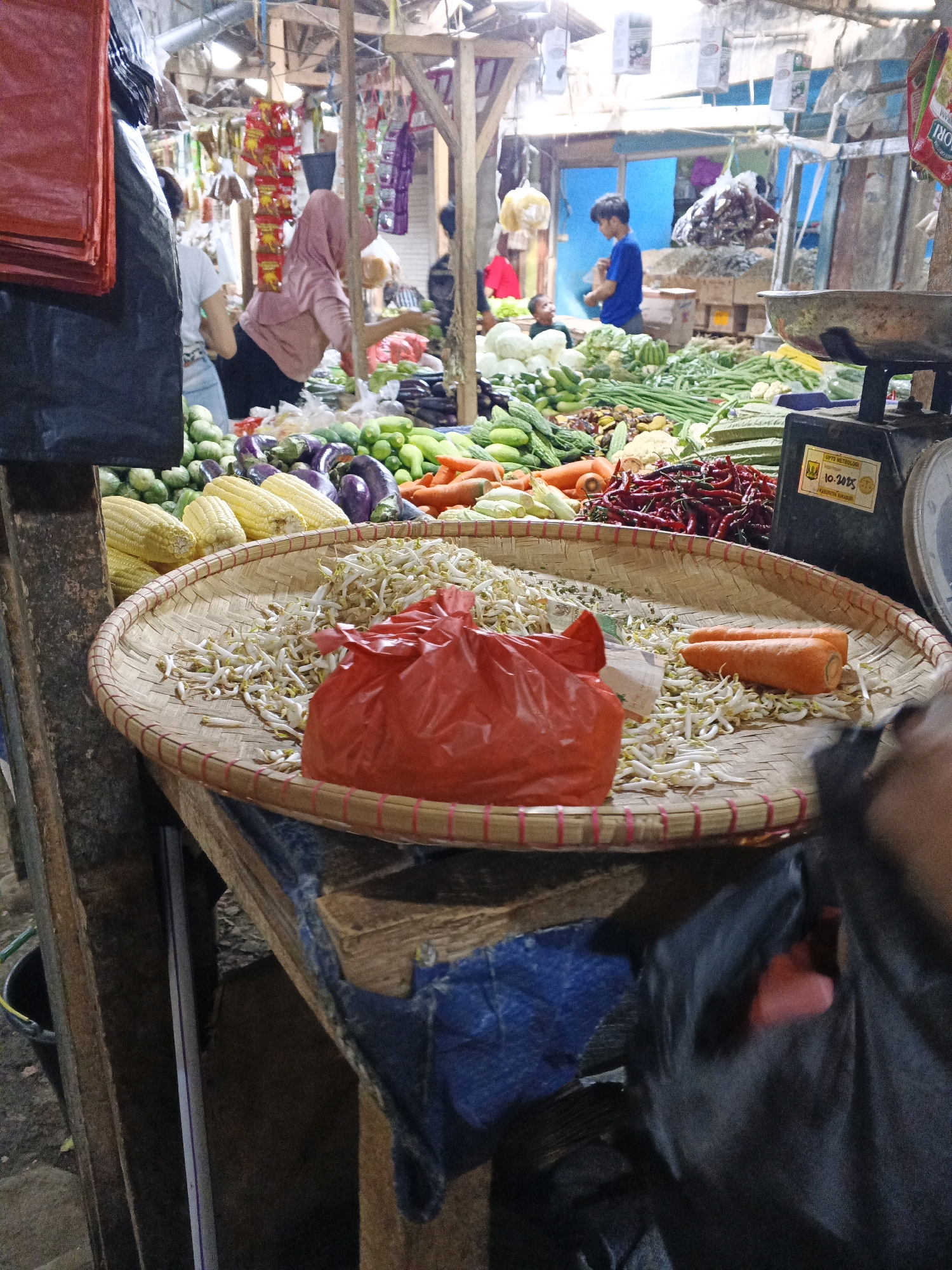 The image depicts a bustling market scene in Warungkiara, Indonesia. In the foreground, a woven tray sits on a wooden table, filled with bean sprouts and a red plastic bag, with two carrots lying on the sprouts. Behind the tray, a variety of vegetables are displayed, including corn on the cob, eggplants, cucumbers, and red chilies. To the right, a black scale with a yellow sticker is visible. In the background, people are seen shopping and interacting at various stalls, with a blue tarp in the distance. The market appears to be outdoors, with wooden structures and hanging goods contributing to the lively atmosphere.