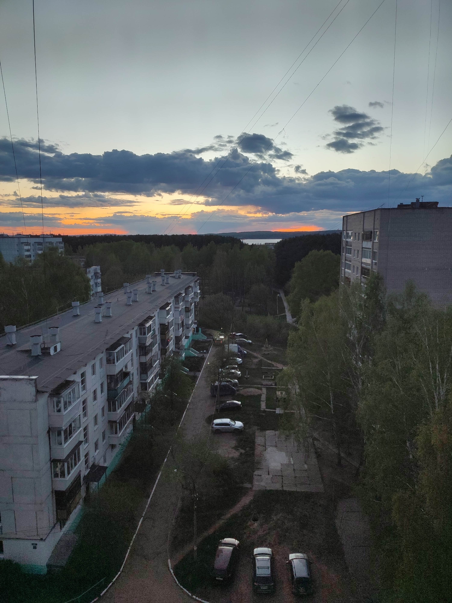 The image depicts an urban landscape at twilight, likely taken from a high vantage point. Two multi-story residential buildings flank a road and wooded area. Several parked cars are visible. The sky is filled with clouds, displaying hues of orange and grey from the setting sun. Power lines crisscross the upper portion of the image. A body of water is visible on the horizon between the trees. The location is unknown, but appears to be a residential area with dense tree cover.