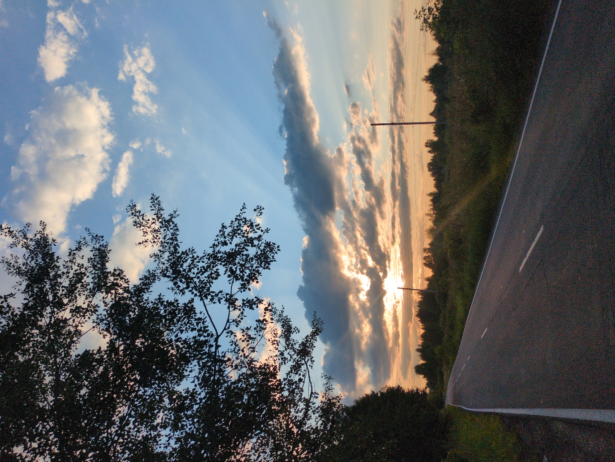 The image depicts a paved road with a white dividing line in the foreground, leading into a landscape at sunset. A dense line of trees forms the horizon. In the sky, the sun is partially obscured by clouds, casting rays of light. There are power lines visible across the horizon, and a single utility pole stands in the middle distance. On the left side of the road, foliage and tree branches overhang the scene. The lighting suggests a late afternoon or early evening setting. The location is identified as Kulebaki, Russia.
