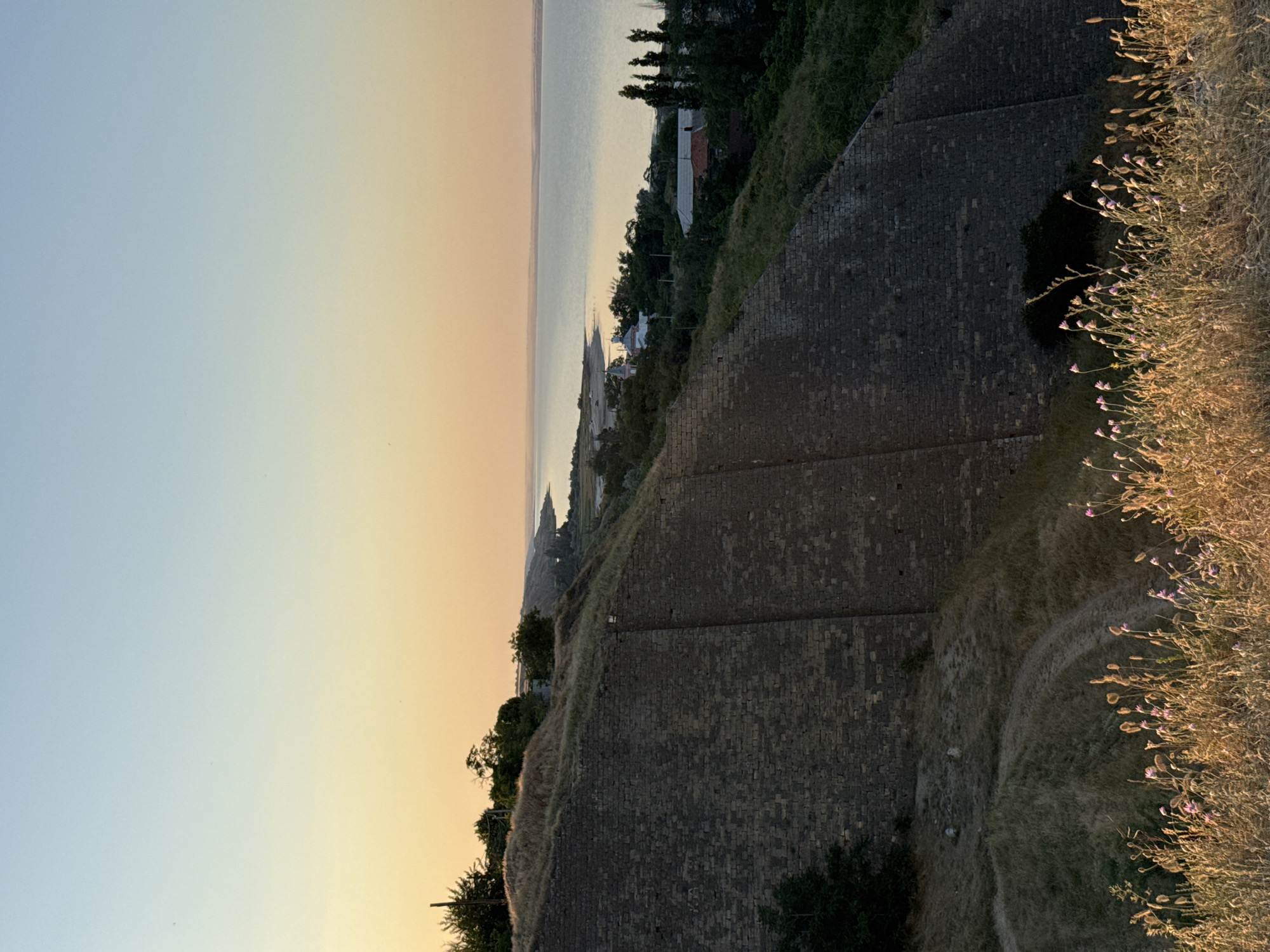 This image depicts a scenic view from atop a stone fortification, likely a historical landmark, overlooking a body of water. The sun is setting, casting a warm, soft glow across the sky, transitioning from a clear pale blue at the top to a soft orange and yellow near the horizon.
The dominant feature in the foreground is the weathered, grey stone wall of the fortification. Its rough-hewn blocks are clearly visible, suggesting age and historical significance. A dry, grassy embankment slopes down from the wall, dotted with sparse vegetation and a few small purple wildflowers. A winding dirt path is visible on this embankment.
Beyond the fortification, a calm body of water stretches towards the horizon. Along its edge, a strip of land is visible with scattered trees and the rooftops of a few buildings, suggesting a coastal settlement or town. The water's surface reflects the soft hues of the sunset.
The overall mood is peaceful and contemplative, with the natural beauty of the sunset and the serene waters. The time of day is clearly sunset, indicated by the warm light and the gradient in the sky. The location appears to be coastal, with the vast expanse of water and the historical structure suggesting a place with a rich past. No people or animals are visible in the image. There is no visible text.