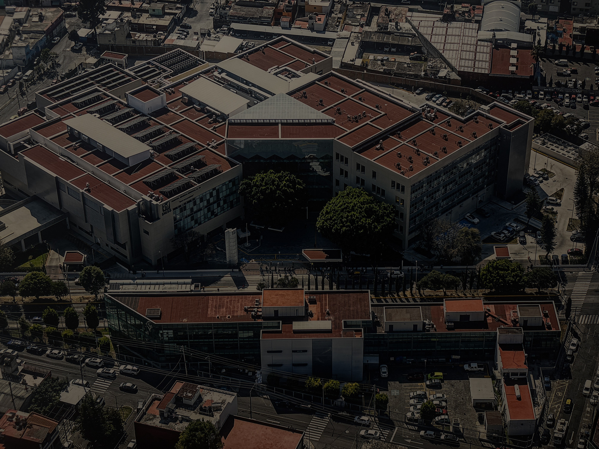 This is an aerial photograph captured during the daytime, likely late morning or early afternoon, under clear skies. The scene depicts a large, sprawling urban complex, confirmed to be the Hospital General Regional IMSS "San Alejandro" in Puebla, Mexico, as indicated by the text and IMSS logo visible on the building's facade.
The main subject is a large hospital building, characterized by its complex, multi-winged structure and numerous interconnected sections. The roofs are predominantly a reddish-brown color, with several flat sections housing various white and grey rooftop units, likely HVAC systems, and what appear to be arrays of solar panels on multiple sections, particularly noticeable in the upper left portion of the complex. A prominent architectural feature is a large, clear pyramidal glass skylight or roof structure located centrally within the main building's layout.
Surrounding the hospital, there's an active urban environment. Streets lined with parked and moving cars traverse the edges of the complex. Multiple parking lots are visible, some filled with vehicles, suggesting a busy facility. Interspersed among the buildings and along the streets are numerous green trees, providing patches of natural vegetation within the urban setting. Smaller, residential or commercial buildings with similar reddish-brown roofs can be seen in the surrounding blocks. Crosswalks and street markings are visible on the roads. No specific event or unusual activity is taking place; the scene portrays a typical bustling day in a hospital and its surrounding urban area.