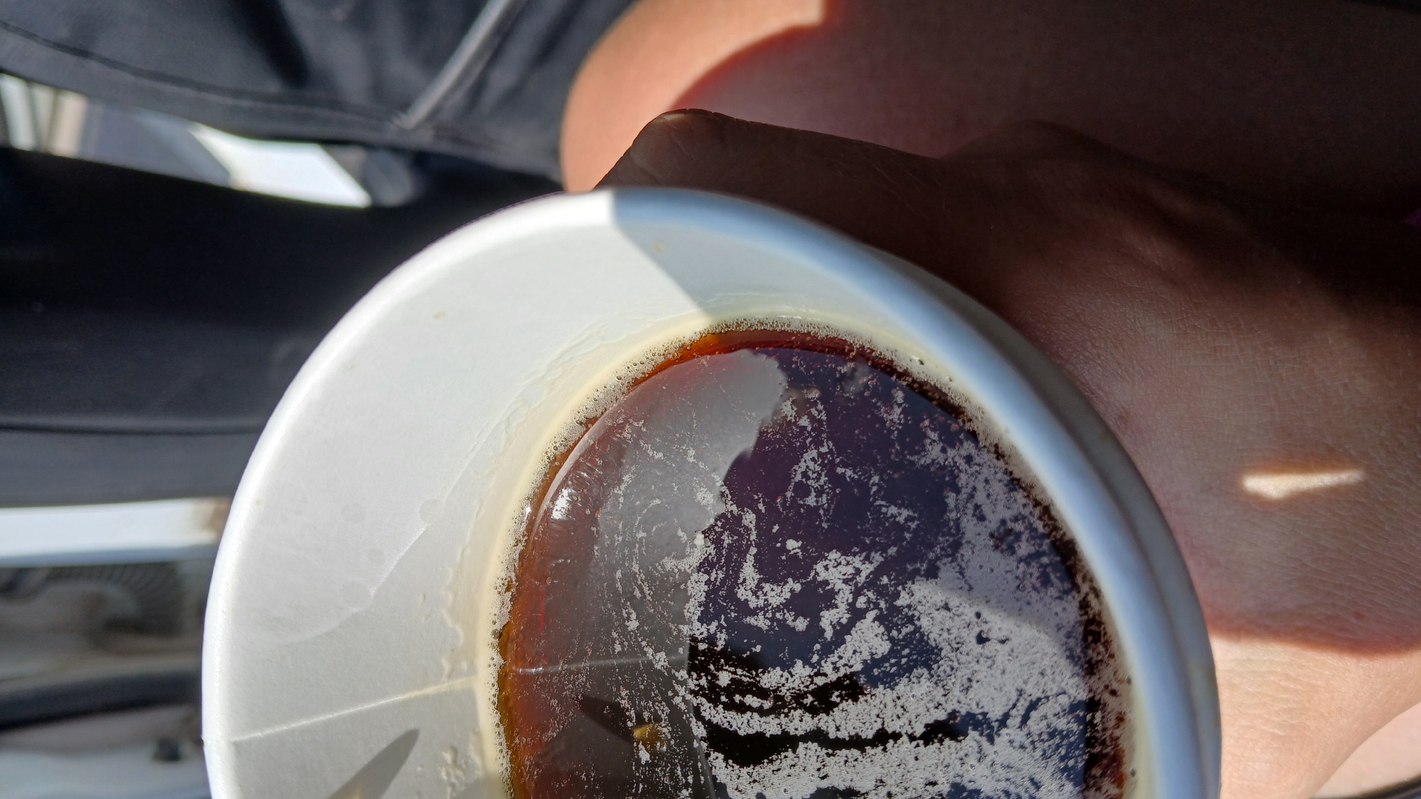 A close-up, overhead shot shows a white paper cup filled with a dark liquid, possibly coffee, with foam on top. The cup is held by a person's hand and arm, which are visible on the right side of the frame. The background is blurred and appears to be outdoors, with sunlight casting shadows on the cup and hand. The environment suggests a casual setting, possibly a park or street.