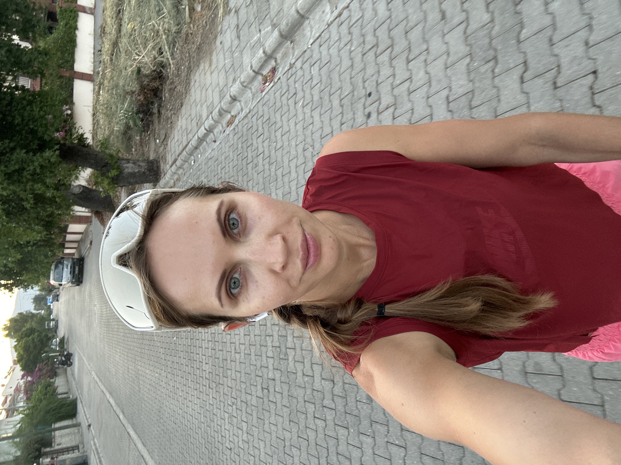 A woman with light brown hair, styled in a braid, and blue eyes is shown in a selfie. She is wearing a sleeveless red shirt, pink shorts, and white sports goggles pushed up onto her forehead. She has white wireless earbuds in her ears. The woman is standing on a cobblestone sidewalk next to a street in Fethiye, Turkey. The street is lined with trees and buildings. Several vehicles, including a van and a motorcycle, are visible on the street in the background. The scene appears to be outdoors during daylight hours.