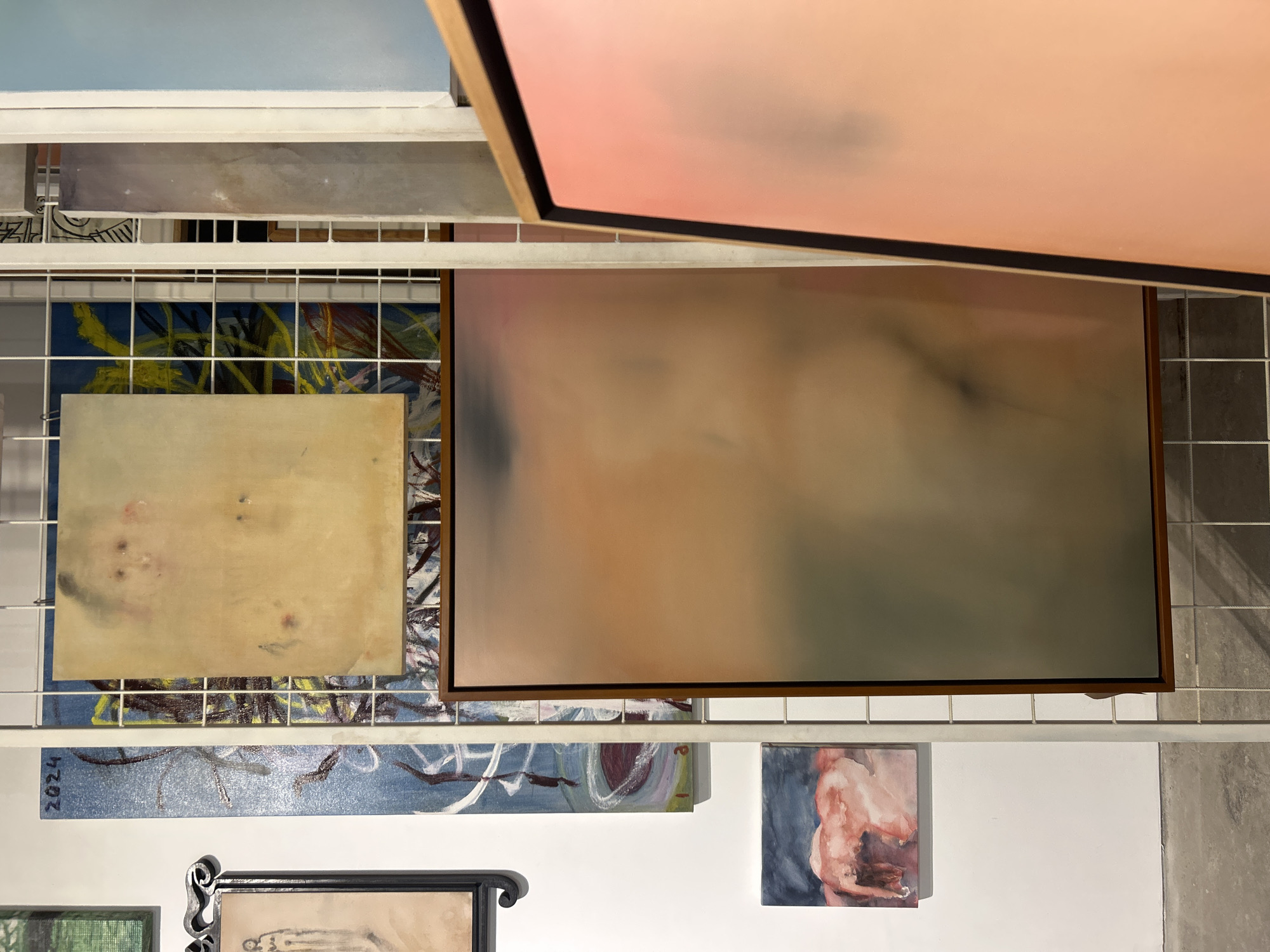 An interior shot depicts a collection of diverse artworks displayed or stored on white metal grids, likely in an art studio or gallery in Mengwi, Indonesia. The overall scene is bright with diffused lighting.
Numerous paintings are visible, showcasing a mix of styles and subjects. In the upper part of the image, a large abstract painting features soft pink and peach gradient tones, partially overlapping another large, muted abstract piece with brown, gray, and faint peach hues, encased in a wooden frame. Below these, a smaller, light yellowish-cream abstract painting with subtle markings rests on a grid. Behind this, another abstract artwork displays vibrant yellow and blue strokes.
Towards the bottom-left, a horizontal blue painting with white and dark linear strokes is visible, bearing the text "2024" on its left edge. Adjacent to it, a smaller, figurative painting depicts a human torso in shades of red, brown, and blue. A black, ornate framed artwork, possibly a drawing, is partially seen at the bottom-center-left, next to a small green textured piece. White walls form the background, with a dark grey tiled surface visible on the far right. The arrangement suggests a layered and somewhat dense display of artistic creations.