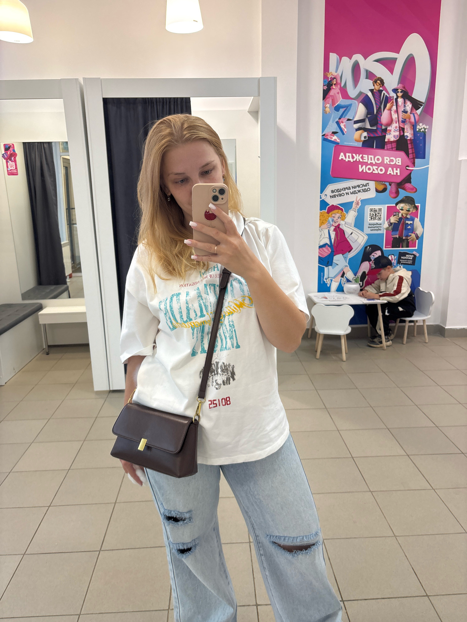 In a brightly lit retail store, possibly a fitting room area in Yuryatino, Russia, a young woman with blonde hair takes a selfie. She is wearing a white t-shirt with a graphic print and light-wash ripped jeans. A dark brown crossbody bag with a gold clasp hangs from her shoulder. In the background, there are changing rooms with dark curtains and benches. To the right, a large promotional banner featuring stylized cartoon characters and Cyrillic text is displayed. Below the banner, a small child sits at a table with two white chairs, appearing to be occupied with an activity. The floor is tiled in a light color.