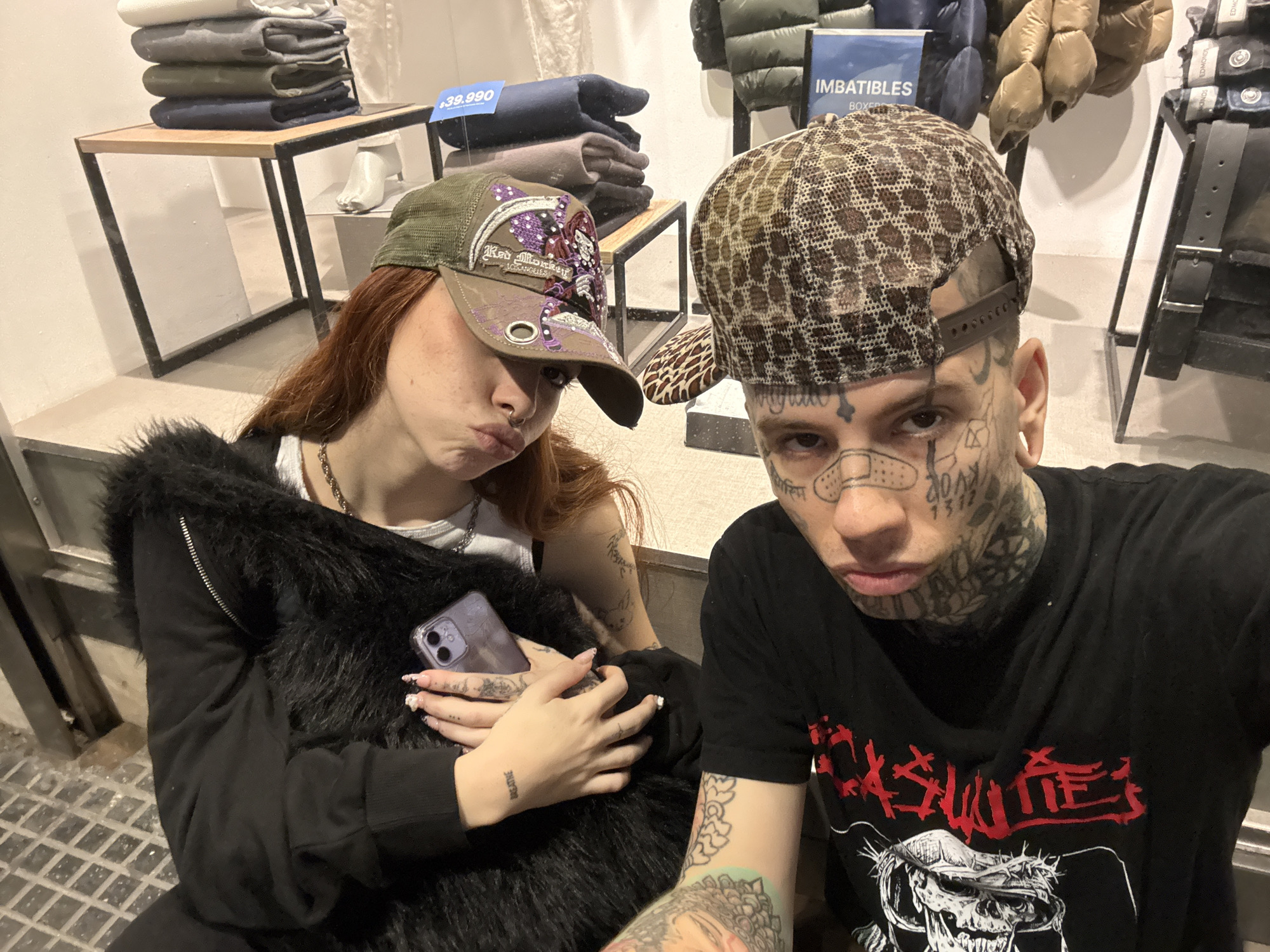 Two people are photographed in what appears to be a retail environment. A woman with red hair and a green cap embellished with purple designs is on the left. She is wearing a black garment with a fur-like texture around the collar. She holds a smartphone in her hands, which are adorned with tattoos. Her facial expression is a pout. A man with extensive face and neck tattoos and a patterned hat stands to the right of the woman. He is wearing a black t-shirt with a red and white design. In the background, there are folded articles of clothing on shelves, and informational signs. The location appears to be indoors, possibly a clothing store. The location in the city of Buenos Aires, Argentina, cannot be confirmed based solely on the image.