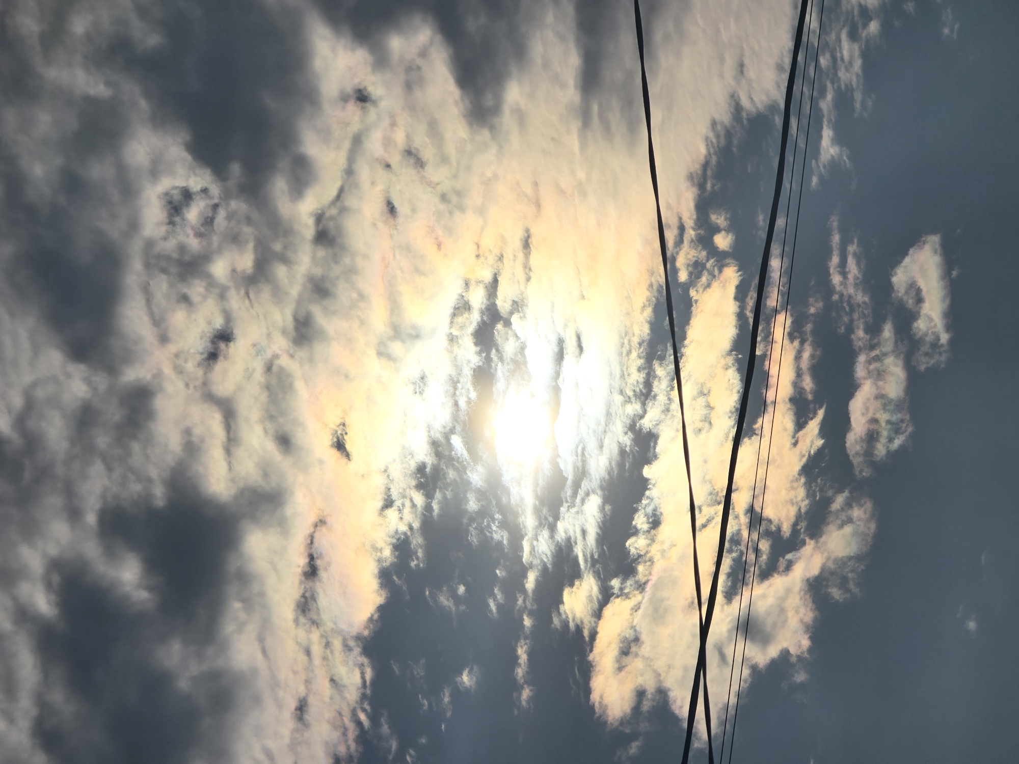 The image depicts a sky with dramatic clouds, illuminated by the sun. The sun is partially obscured by the clouds, creating a bright, hazy glow. The clouds themselves display a range of colors, from dark grays and blues to lighter whites and yellowish-pinks, suggesting the presence of iridescent cloud iridescence. Several dark, thin wires run horizontally across the frame, indicating the presence of utility lines. The lower portion of the image shows a darker, more uniform sky. There are no people or distinct man-made structures visible in this particular shot. The scene is an outdoor sky view, likely from ground level or a slightly elevated position, overlooking the sky above Ермолаево, Russia.