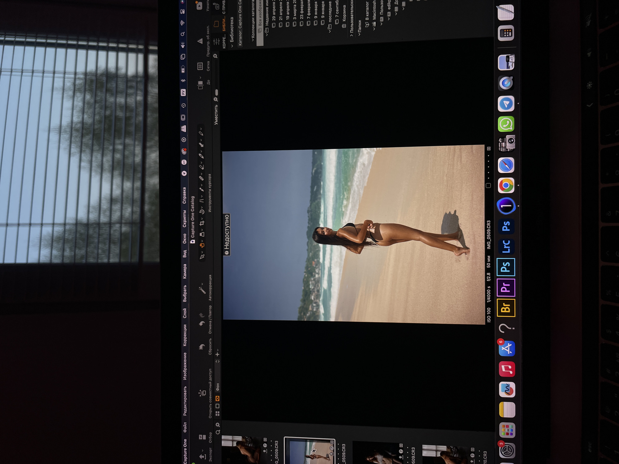 The image shows a MacBook screen displaying the Capture One photo editing software. A photograph of a woman on a beach is open in the software. The software interface includes file management options, editing tools, and metadata information about the photo.
Other icons, presumably of applications and settings, appear in the dock, along the right edge of the screen.
The laptop is situated near a window with closed blinds. A blurred view of trees and the sky is visible outside.