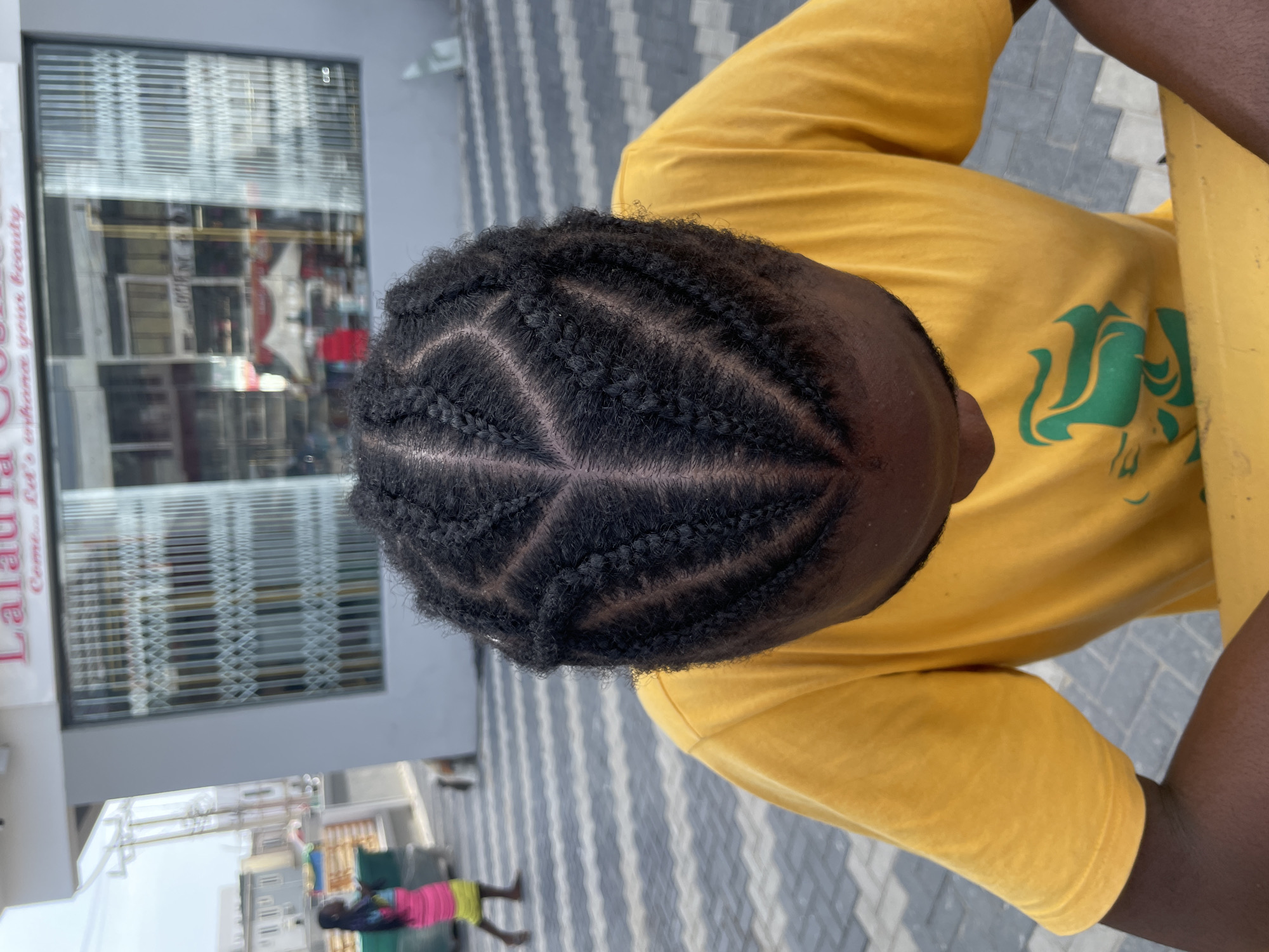 A top-down shot depicts a person with their hair braided into a symmetrical design with a central triangle pointing towards the forehead. The person is wearing a yellow t-shirt with a green logo on the right side of the frame.
The background features a building with a partially closed metal grate over the storefront window. The sign "Lalaurla" is visible on the storefront. A pedestrian is walking on a pavement consisting of white and grey alternating stripes, which extends to the bottom of the image. The setting is Lekki, Nigeria.