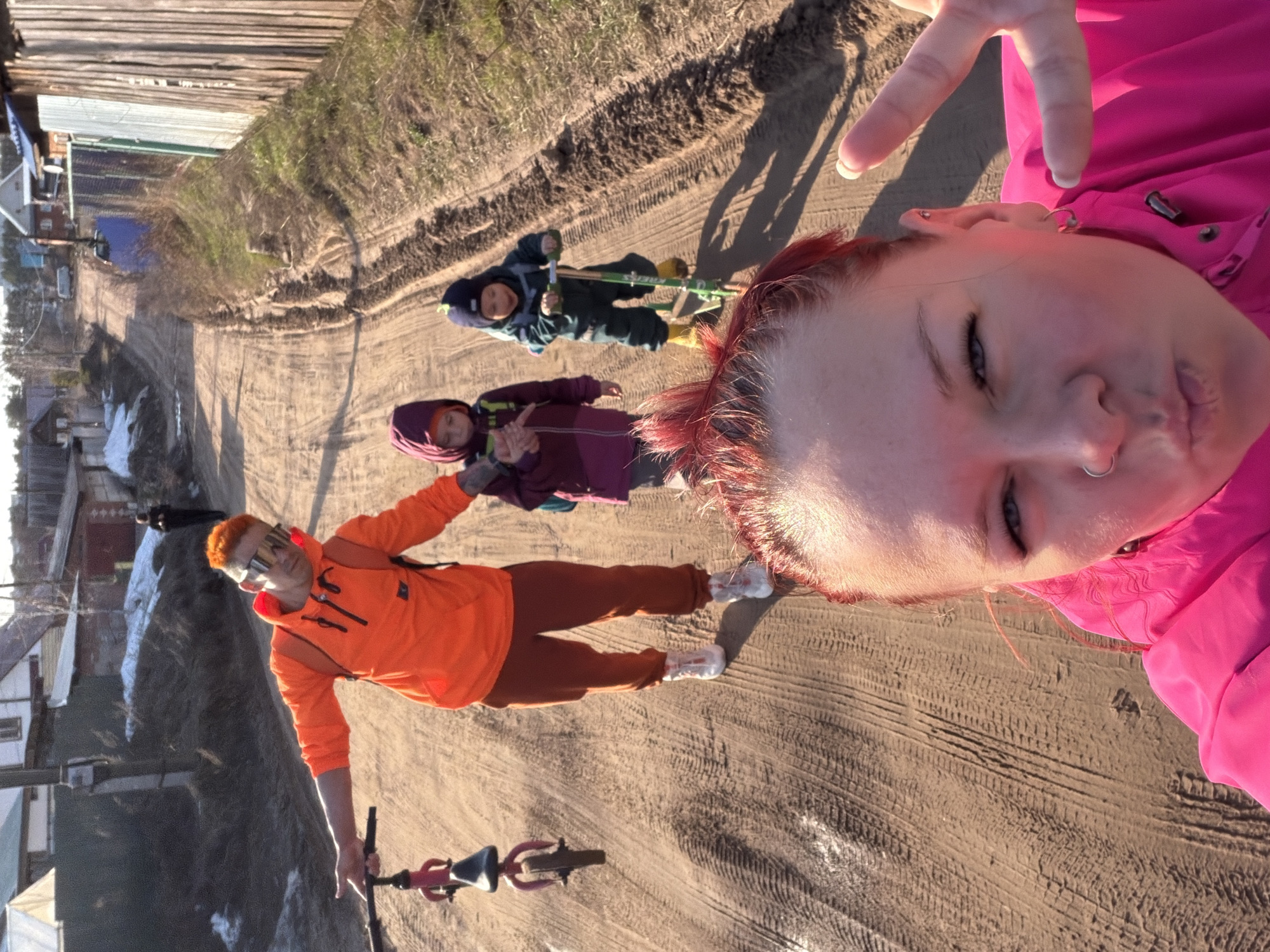 The image depicts a selfie taken in what appears to be a rural or semi-rural setting near the city of Chaikovsky, Russia. A person with red hair and a pink jacket is holding the camera. In the background, there are three children; one is on a pogo stick, another one makes a peace sign, and the third is standing on a bike. The setting is a dirt road with houses and some snow visible on the sides. The lighting suggests it is daytime.