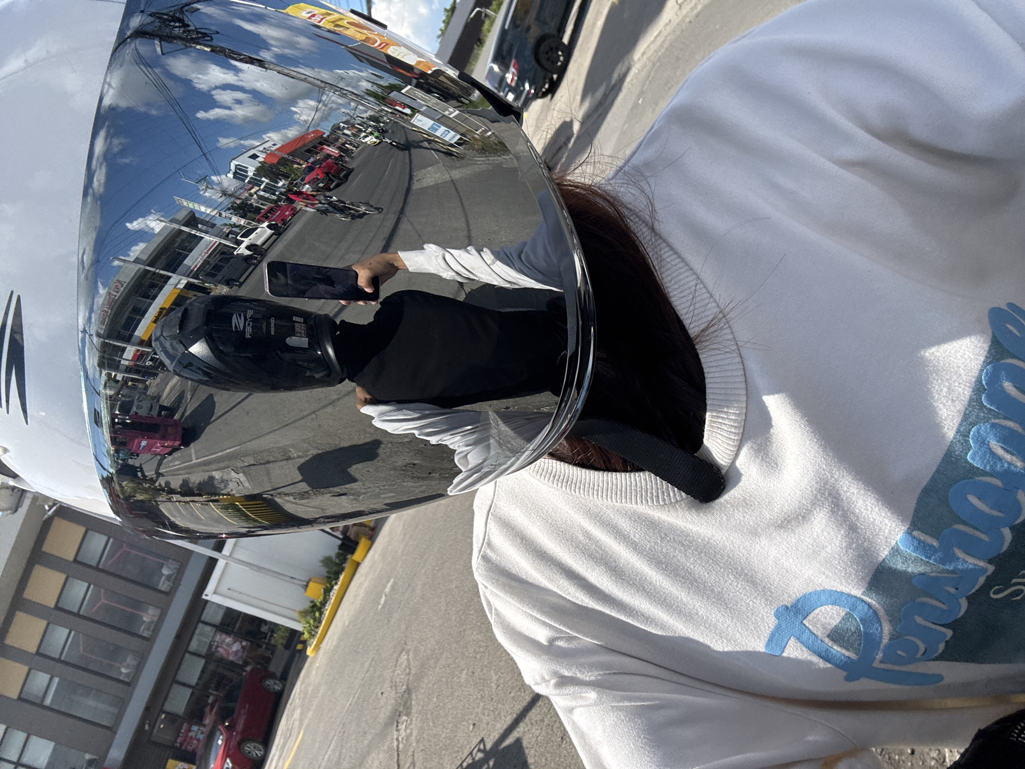 The image shows a first-person perspective of someone wearing a white long-sleeved shirt and a motorcycle helmet with a highly reflective visor. The visor reflects a busy street scene with buildings, vehicles, and a bright blue sky with clouds. The buildings appear to be commercial establishments, with signage visible. Vehicles include cars, motorcycles, and what looks like a pink jeepney. The road is paved, and there are some yellow markings. The presence of a jeepney and the general streetscape suggest an urban environment in the Philippines. Based on the distinctive architecture of the commercial buildings and the commonality of jeepneys and motorcycle riders, this scene is consistent with locations in Davao City, Philippines. The person is holding a smartphone up, presumably to take the photo, with their hand and arm visible in the reflection. The person is wearing a black vest over their shirt.