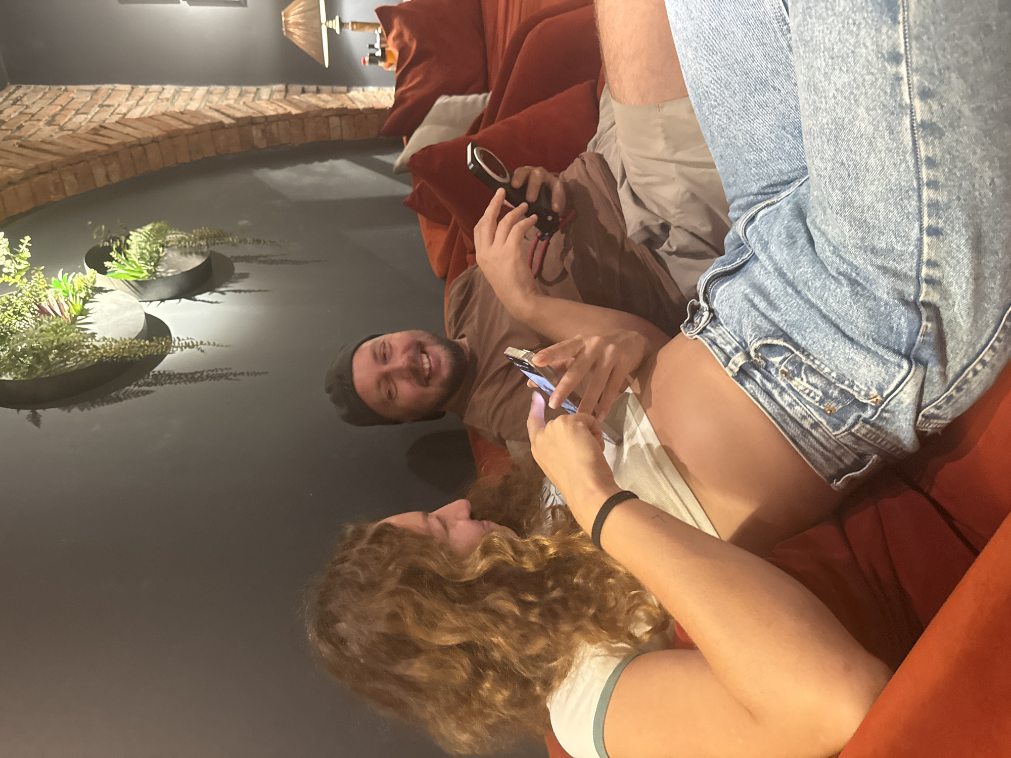 A pregnant woman and a man are sitting on a red couch. The woman is on the left, facing to the right, and is looking at her phone. She is wearing a white t-shirt with a green collar and blue jeans. The man is on the right, facing to the left, and is looking at his phone. He is wearing a brown t-shirt and khaki shorts. There are plants on the wall behind them, and a brick archway to the right. A lamp is visible on a table behind the man. The image appears to have been taken indoors. There is no information to suggest this image was taken in Hà Nội, Vietnam.