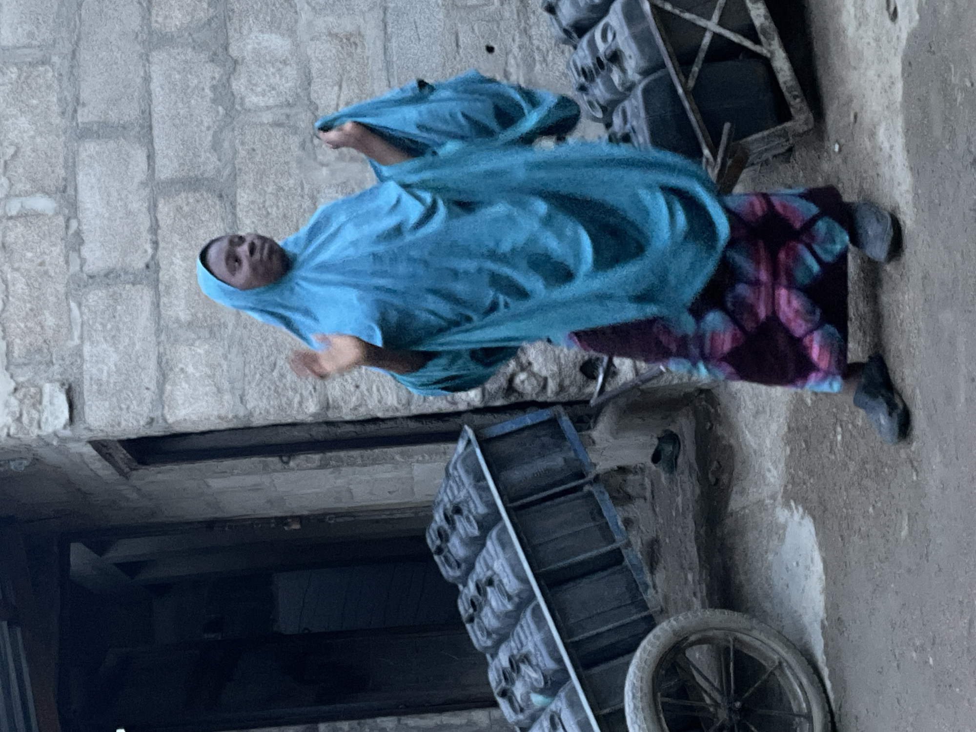 A woman in a blue headscarf and dress is walking in an urban area in Unknown, Unknown. She is facing left in the image. There are buildings in the background and objects on the ground near her.