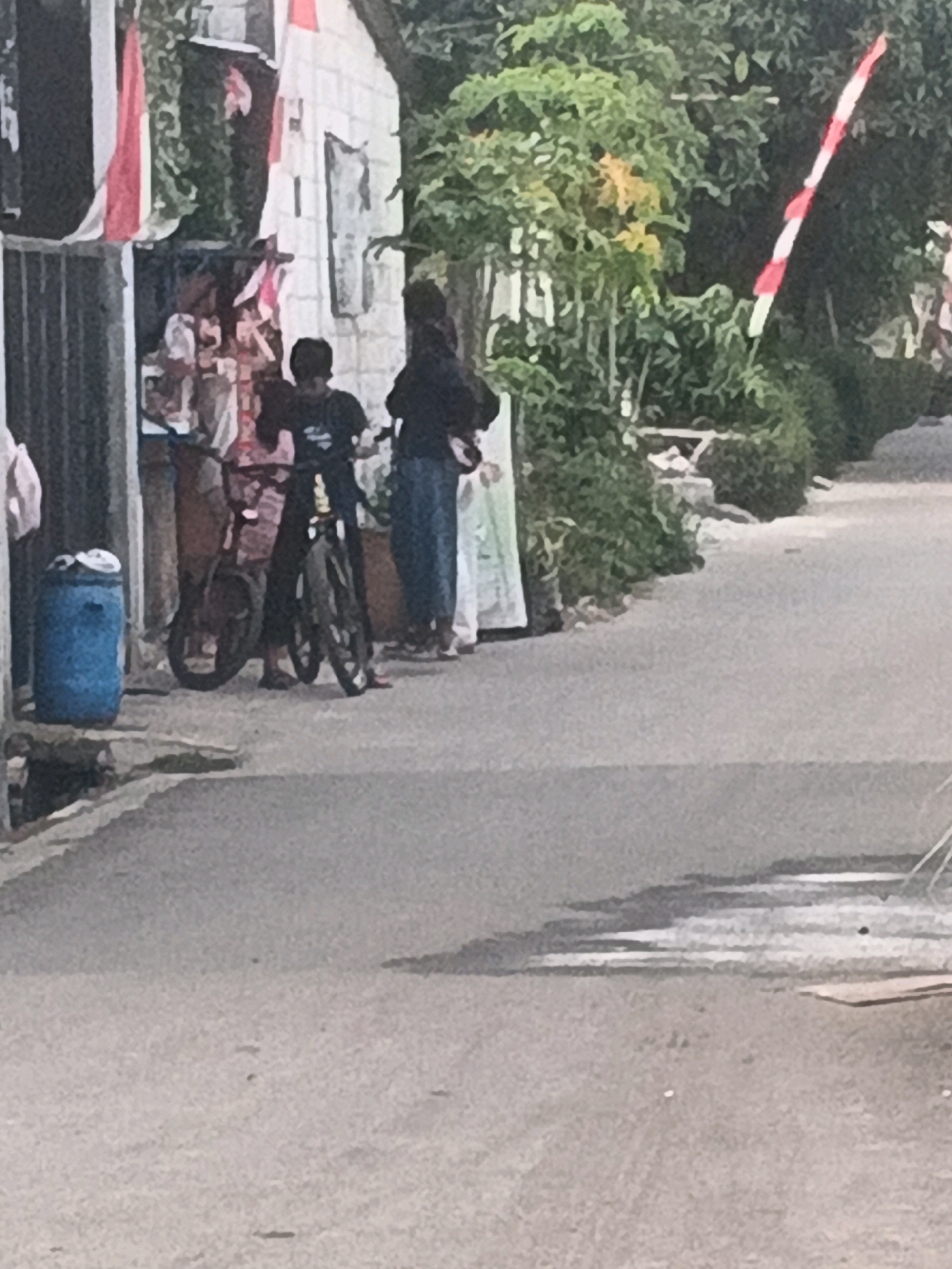 A full shot shows a street scene in Kadu, Indonesia, with a white building on the left and a paved road. Two individuals are visible near the building. One person is wearing a dark shirt and dark pants, standing beside a bicycle. The other person is wearing a dark hooded garment and blue pants, also near the bicycle. A blue barrel is situated to the left of the building. Indonesian flags are displayed on the building and a pole on the right side of the frame. The environment is outdoors, with trees and foliage visible behind the building and on the right. The road appears to be a street with a crosswalk marking.
