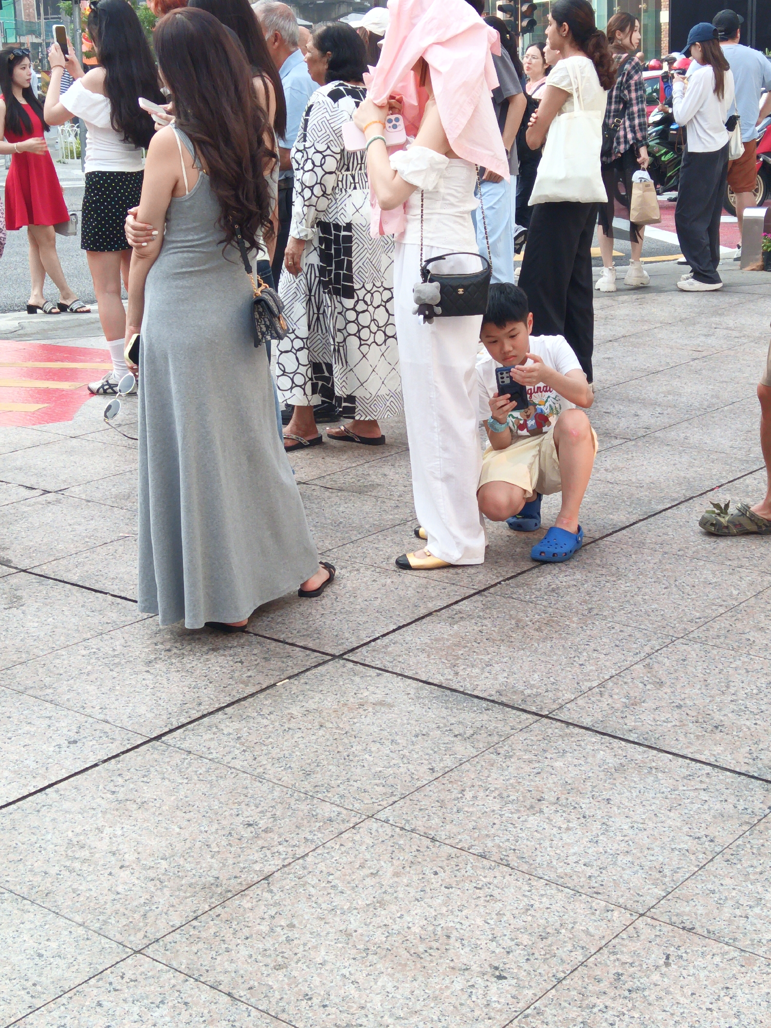 The image depicts a street scene with several people gathered on a paved walkway. In the foreground, a young boy is crouching and looking at his phone. He is wearing a white t-shirt and yellow shorts, with blue Crocs on his feet. Behind him, a woman in a long grey dress is standing, holding a purse. Further back, a group of people are standing around, some holding phones. A woman in a pink headscarf is also visible. The environment appears to be an urban setting, with buildings and possibly shops in the background, although they are out of focus. The ground is a patterned tile. The presence of a young boy engrossed in his phone and other individuals using their devices suggests a typical urban scene where technology is prevalent.