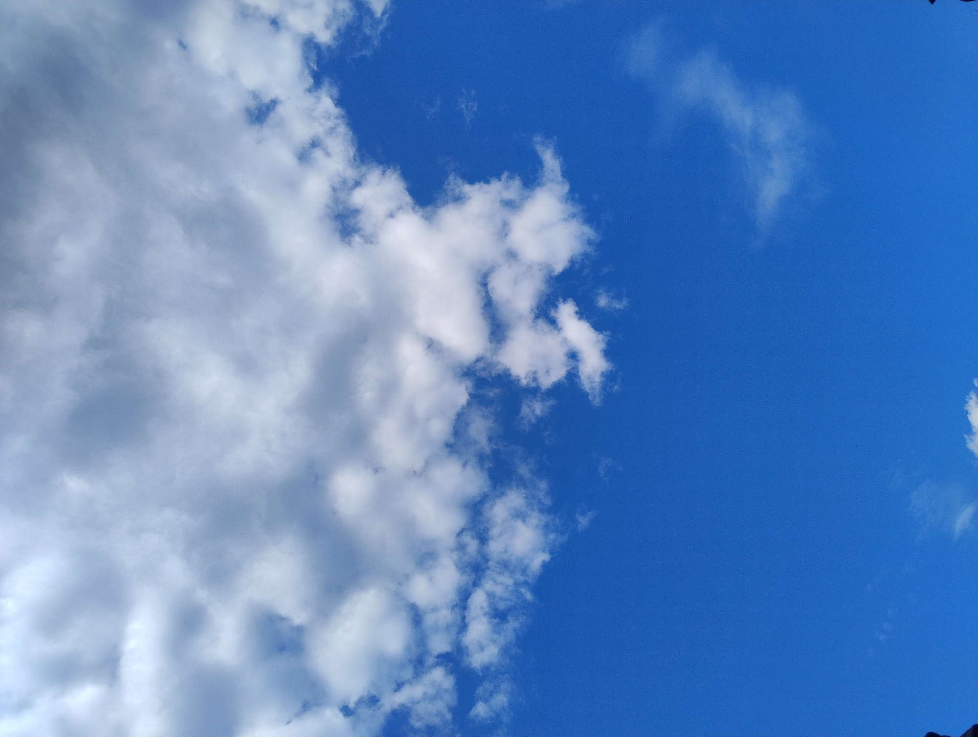 The image displays a vibrant blue sky filled with white clouds. The clouds are dense and fluffy, occupying the top half of the frame, with some thinner, wispy clouds scattered across the lower portion. The overall impression is one of a clear and bright day. No people, subjects, objects, actions, or interactions are visible. The location is identified as Unknown, Unknown.