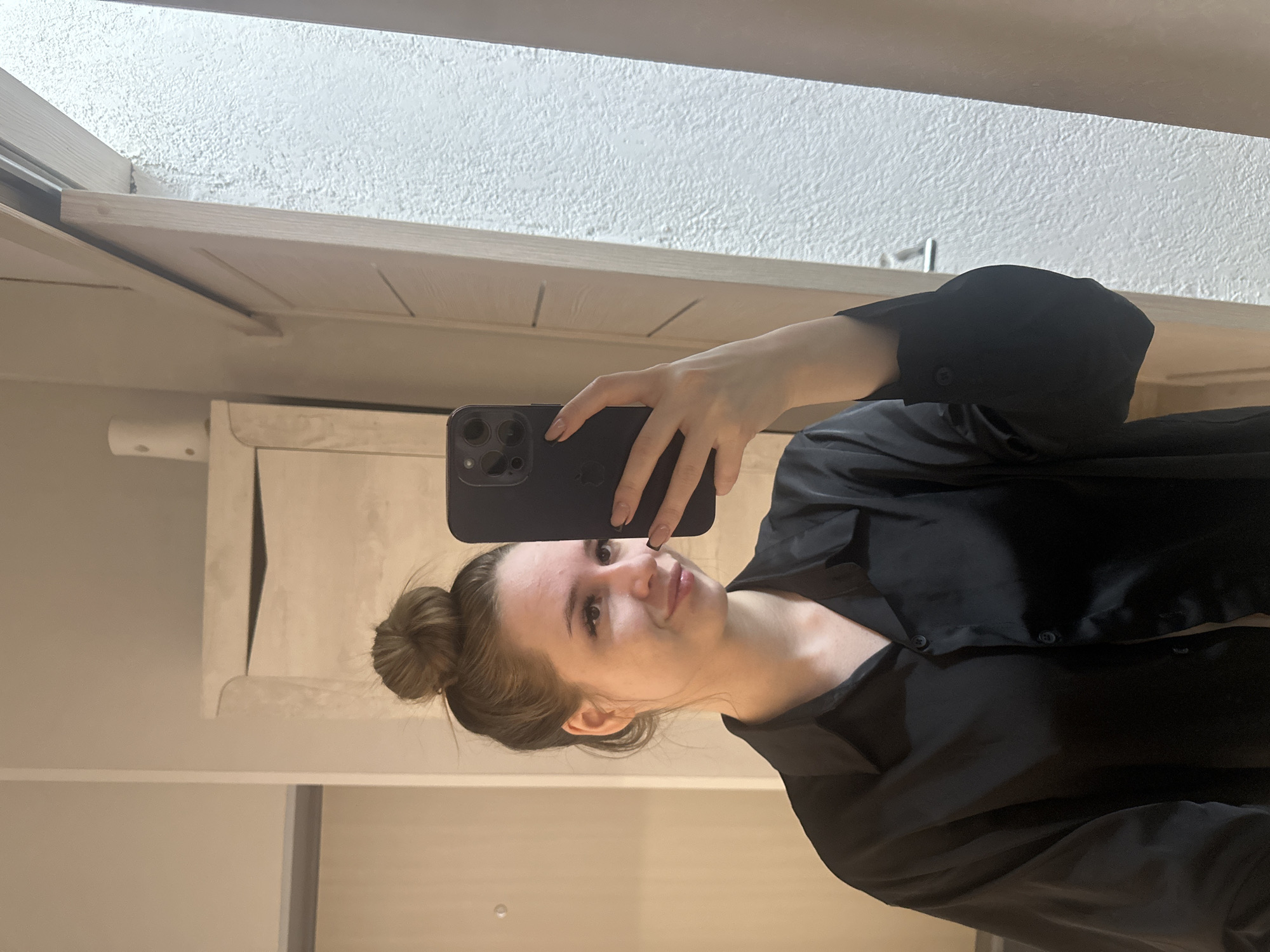 A person is holding a phone to take a selfie in an indoor location. The individual has fair skin, hair pulled back in a bun, and is wearing a black collared shirt. The phone is black with multiple camera lenses visible. The background consists of neutral-toned walls and trim, with a textured white surface above, possibly a ceiling. A portion of what appears to be a cabinet or built-in fixture is also visible. The location in the city of Unknown, Unknown, is not discernible from the image.