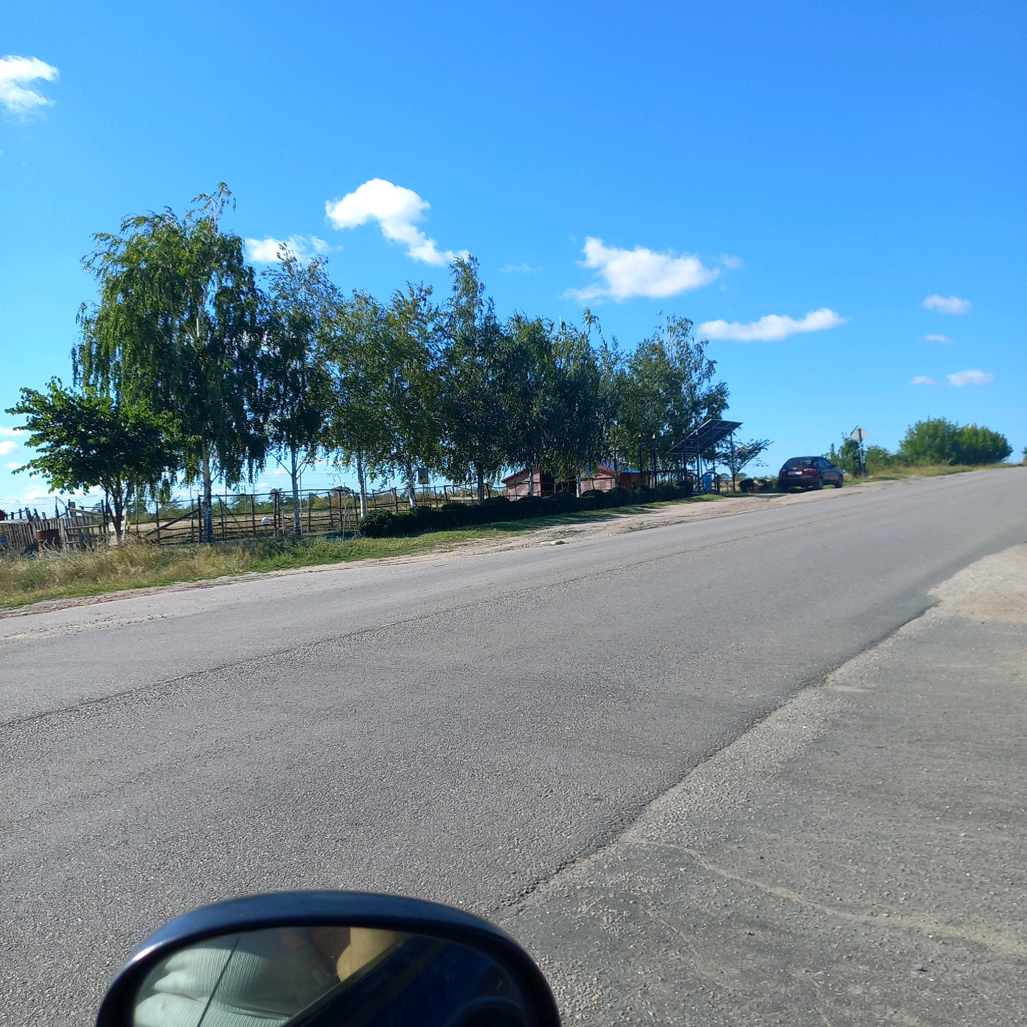 The image depicts a rural road scene on a clear, sunny day. A paved road curves to the right, with a grassy embankment on the right side and trees lining the left side. Behind the trees, there is a wooden fence and some structures, including a small building and what appears to be a solar panel array. A dark-colored car is parked on the side of the road. The sky is bright blue with a few scattered white clouds. The foreground shows the reflection of a car's interior, specifically a car mirror and what looks like a person's arm, in the rearview mirror of the vehicle from which the photo was taken.