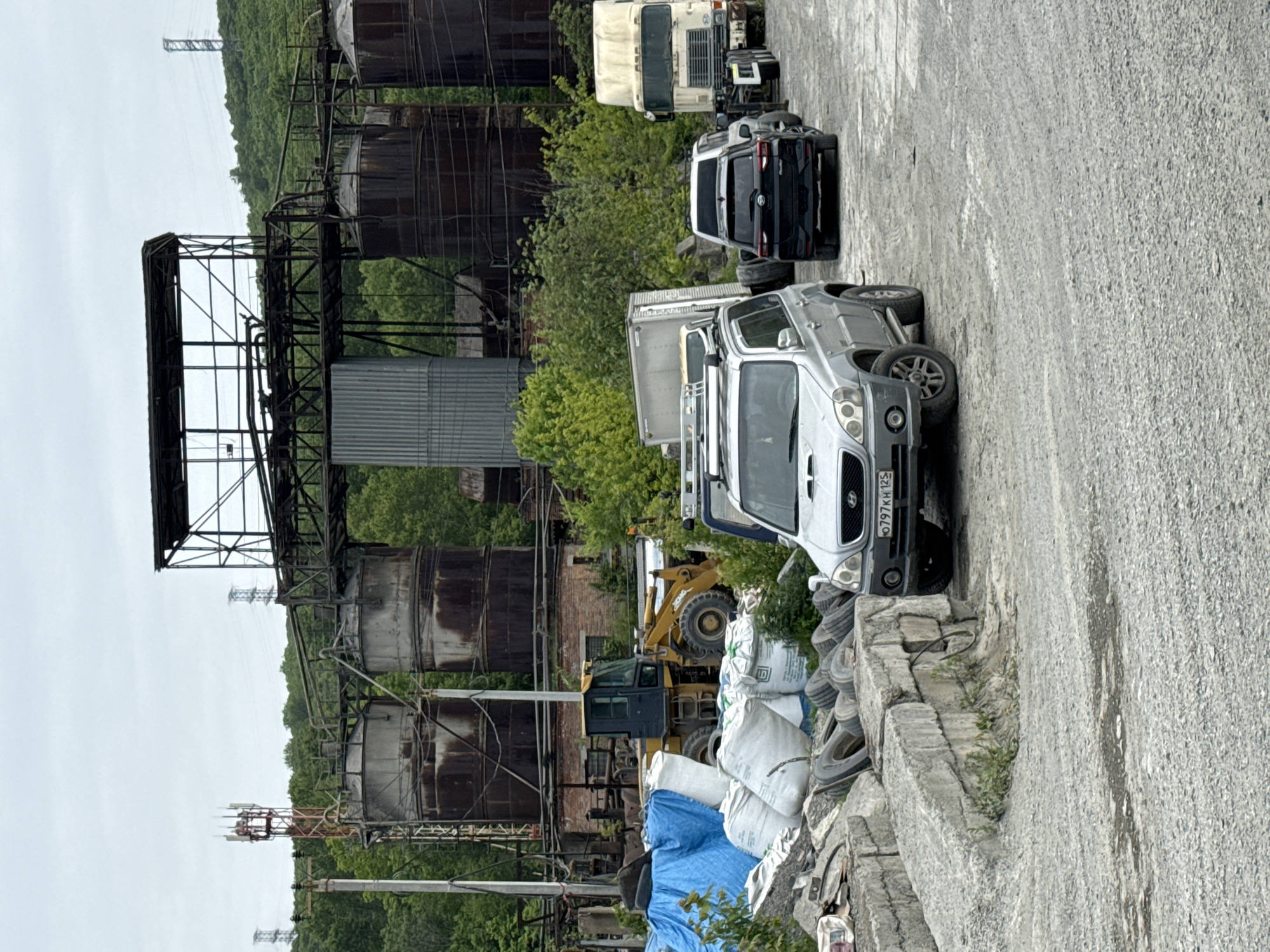 This image depicts an industrial or storage yard scene, likely in an abandoned or disused state, with a backdrop of a forested hill under an overcast sky. No people are visible.
The scene is dominated by several vehicles and industrial structures. In the foreground, a silver SUV-like vehicle (Hyundai Terracan) is parked, showing a Russian license plate "о 797 KH 125". The "125" regional code corresponds to Primorsky Krai, confirming the location context of Vladivostok, Russia. Adjacent to it are piles of used tires and stacked concrete blocks. Further back, a yellow front-end loader (appearing to be an XGMA model, though text is partially obscured) is visible, surrounded by white sacks and a blue tarp.
Behind these, multiple large, heavily rusted industrial tanks are prominent, interconnected by pipes and metal scaffolding. A corrugated metal structure stands vertically near them. In the mid-ground, a black SUV and a white box truck or large van are parked. Further back, a large white truck chassis, possibly a semi-truck cab without its trailer, is visible.
In the distance, an electricity pylon is seen on the hill, and closer to the industrial structures, a cellular tower with antennas is present. The ground is a rough, paved surface. The overall impression is one of an old industrial area, possibly undergoing demolition or serving as a salvage/storage yard, with machinery and vehicles present but no discernible activity taking place. The time of day appears to be daytime, with an overcast sky.