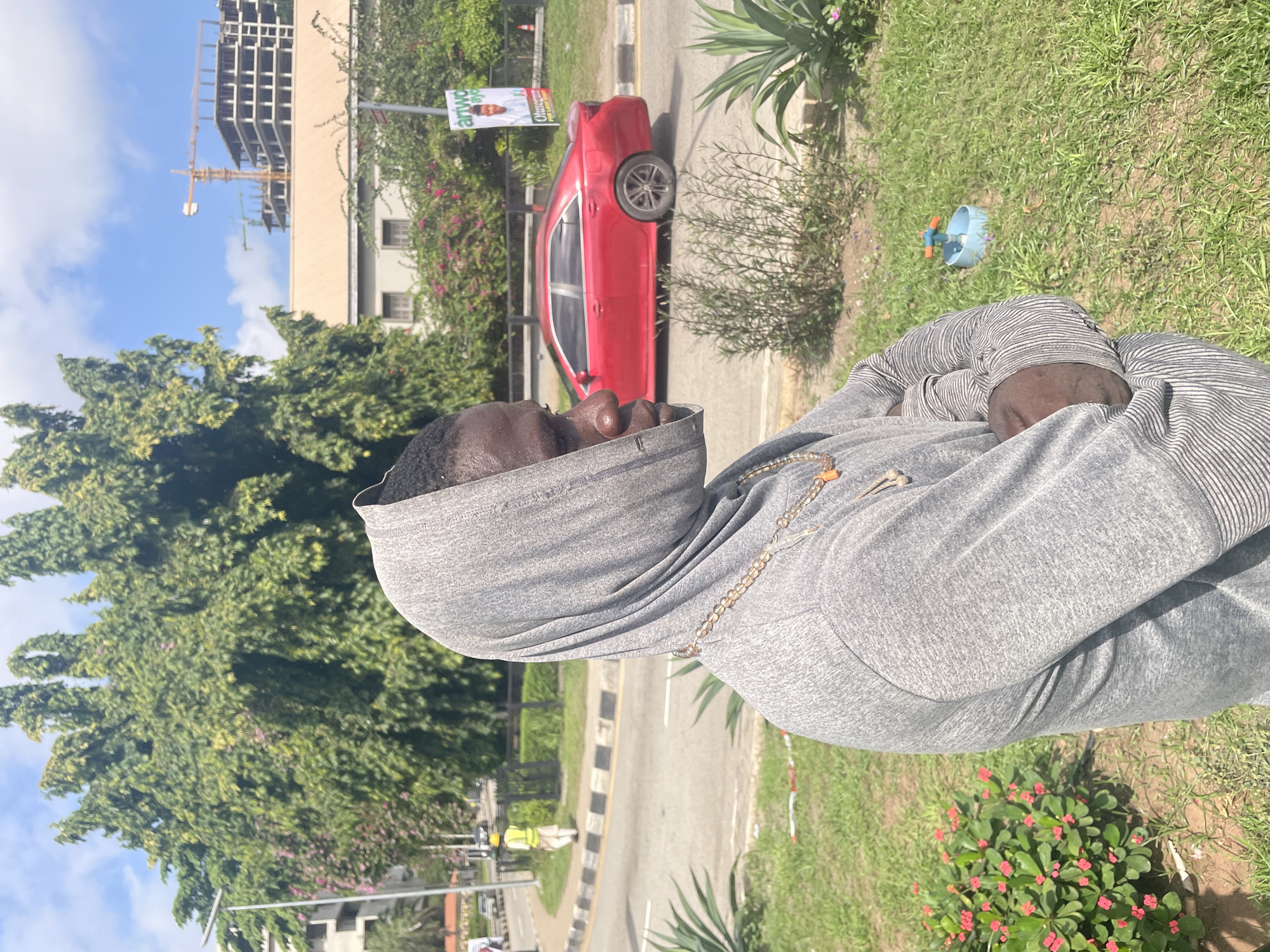 A person wearing a grey hoodie is standing outside on a sunny day. A red sedan is parked on the side of the road. Construction can be seen in the background. The location is Ikoyi, Nigeria.