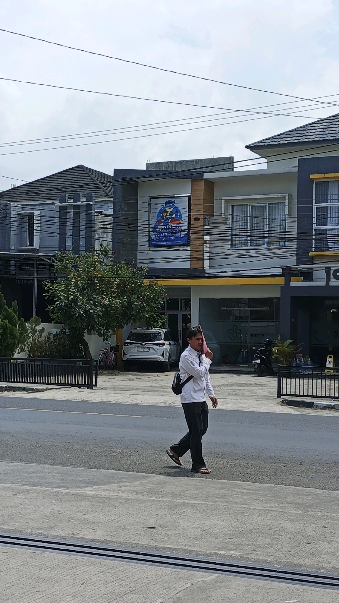 A man wearing a white long-sleeved shirt and dark pants walks across a paved road in front of a row of buildings. He is holding a pink cloth to his face, possibly to wipe sweat or shield himself from the sun. A dark bag is slung across his shoulder. In the background, there are several modern buildings with dark gray and yellow accents. A white SUV is parked in front of one of the buildings, and a bicycle is leaning against a tree. Overhead, power lines crisscross the sky, which is overcast. The scene appears to be a commercial or residential area in Pangandaran, Indonesia, indicated by the signage and architectural style typical of the region.