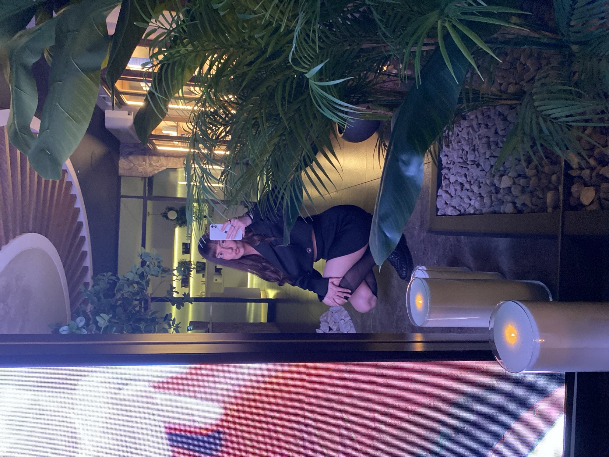 A person, presumably female, is kneeling to take a selfie with a mobile phone. The person is wearing a black outfit with black stockings and shoes. The surroundings appear to be a commercial or public setting with greenery, decorative stones, and lighting elements like candles in glass containers. The setting could be an outdoor patio or a restaurant area. There is a wall with a patterned design and other plants visible in the background. The image is cropped near the bottom, obscuring details of what appears to be a large screen display. There is no verifiable information to locate this specific scene within the city of Votkinsk, Russia.