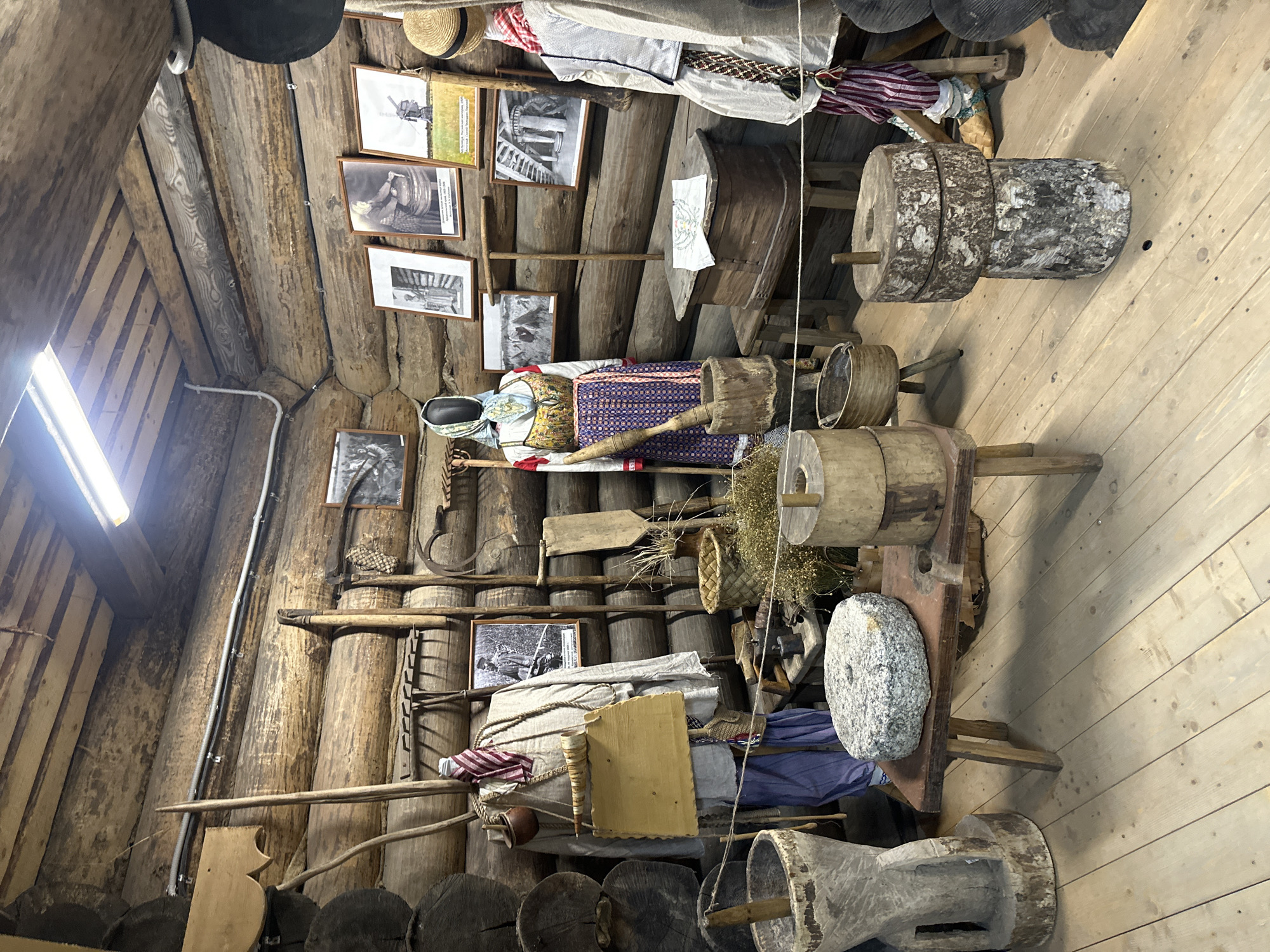 This image depicts an interior scene within a rustic, log-built structure, likely a museum exhibit showcasing traditional Russian rural life, located in Nevskii Parkleskhoz, Russia. The walls and ceiling are constructed from thick, rough-hewn logs. The floor is made of wooden planks.
In the center of the room, a mannequin dressed in traditional Russian folk attire stands. The attire consists of a white blouse with red embroidery, a colorful patterned sarafana (a type of jumper dress), and a headscarf. To the left of this mannequin, another mannequin is visible, partially obscured, also wearing traditional clothing.
Various historical tools and artifacts are displayed throughout the scene. These include wooden agricultural implements such as rakes, scythes, and a shovel leaning against the wall. Several large wooden grinding stones, some appearing to be millstones, are placed on the floor and on a wooden bench. One prominent grinding stone, grey and circular with a hole in the center, rests on a low wooden table. Wooden buckets and baskets are also present.
The walls are adorned with framed photographs depicting scenes of rural life, possibly from the early 20th century. These images show people engaged in agricultural work, as well as architectural elements like windmills and old buildings.
A wooden chest sits on a low stool in the foreground, with a piece of embroidered fabric draped over it. The lighting in the room appears to be artificial, with a long fluorescent light fixture visible on the ceiling. The overall impression is one of a preserved historical exhibit intended to educate visitors about the past.