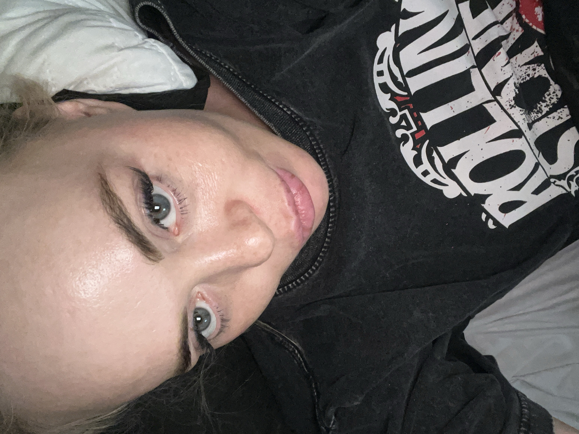 A close-up, overhead, point-of-view shot shows the face of a blonde woman with blue eyes and fair skin. She is wearing a black t-shirt with a Rolling Stones logo. The woman is lying on a white surface, possibly a pillow or bedsheets, and appears to be looking up at the camera. The lighting suggests an indoor setting, and the overall mood is casual.