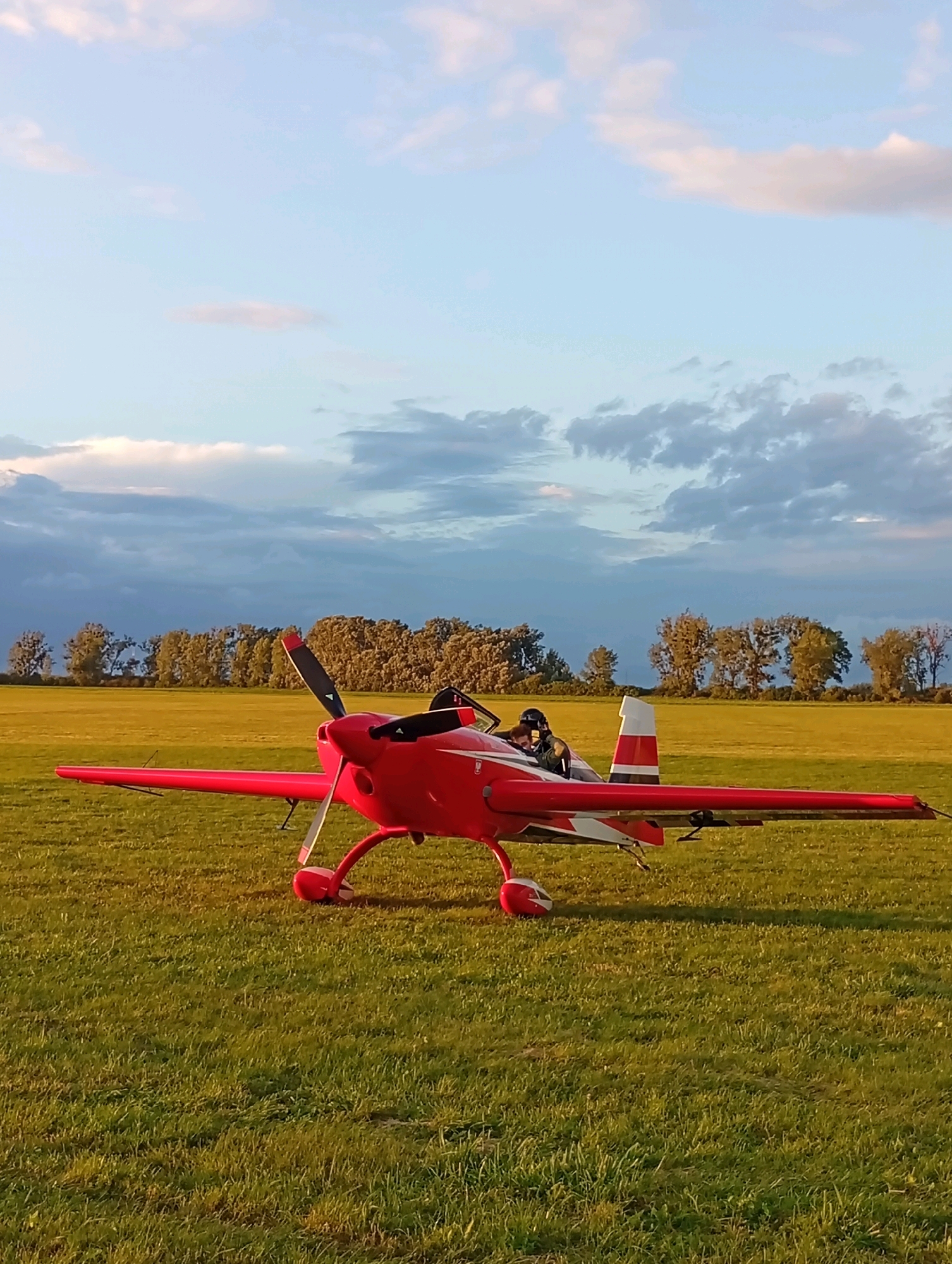 A bright red, single-engine airplane is parked on a grassy field. A person wearing a helmet and dark clothing is seated in the cockpit. The airplane has a propeller at the front and a tail with red and white stripes. In the background, a line of trees forms a distant horizon against a sky with scattered clouds. The scene is captured in Szewce, Poland, suggesting an outdoor, possibly rural or recreational setting for aviation activities. The lighting indicates it might be late afternoon or early evening, with the sun casting long shadows.