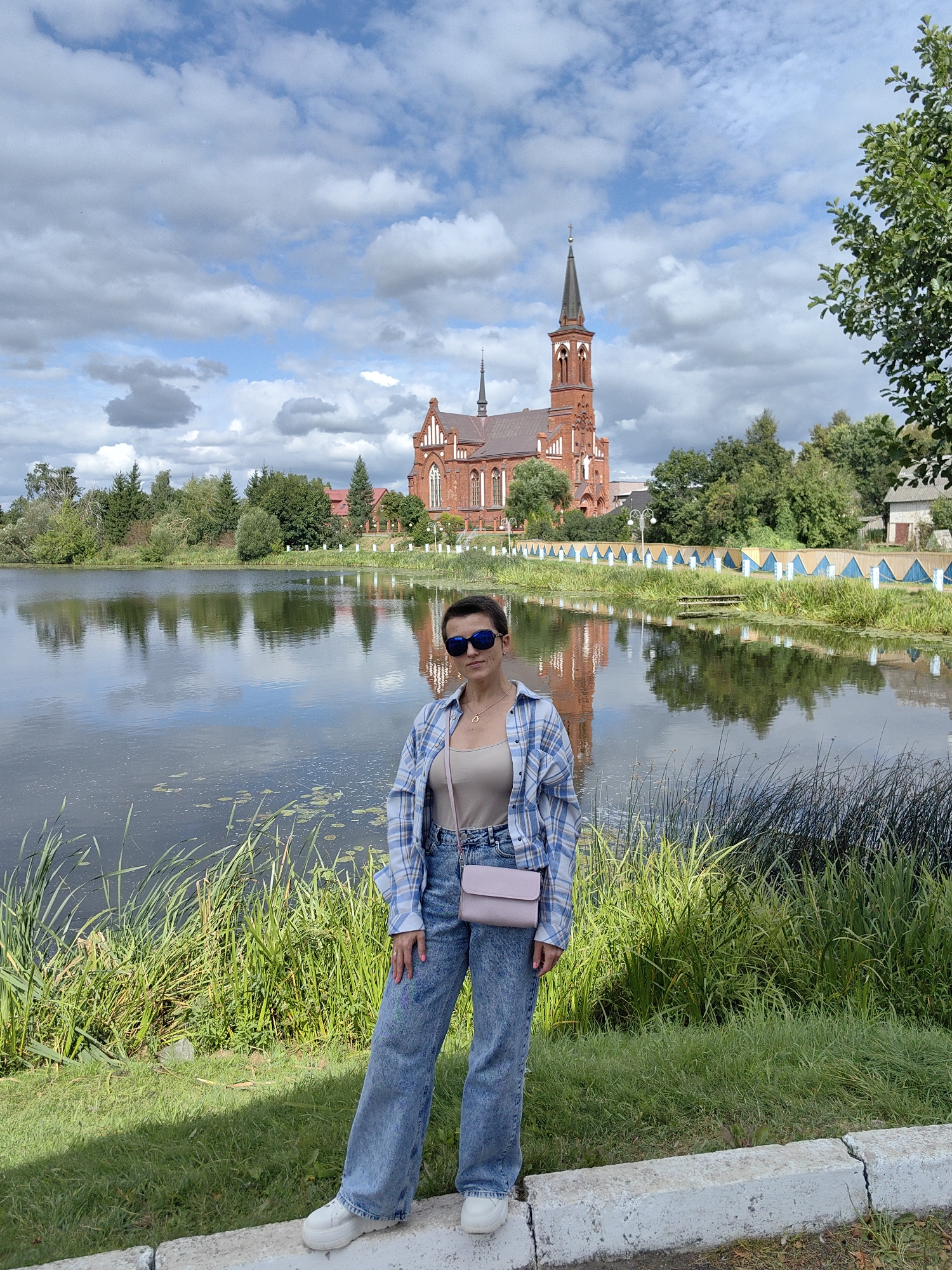 A medium shot captures a woman standing on a grassy embankment beside a tranquil body of water. The woman, with short dark hair and sunglasses, wears a light-colored tank top, an open blue and white checkered shirt, and loose-fitting, light-wash jeans. A pale pink cross-body bag is slung over her shoulder. Her feet are clad in white sneakers. In the background, a large, red brick church with a tall steeple stands prominently, its reflection shimmering on the water's surface. The scene is set in the city of Pastavy, Belarus, indicated by the presence of the notable church and the natural landscape. Lush green reeds and grass line the foreground, contrasting with the calm, reflective water. The sky above is a mix of blue and scattered white clouds, suggesting a partly cloudy day. A decorative fence, with blue and white elements, runs along the far bank of the water, behind which are trees and some buildings.