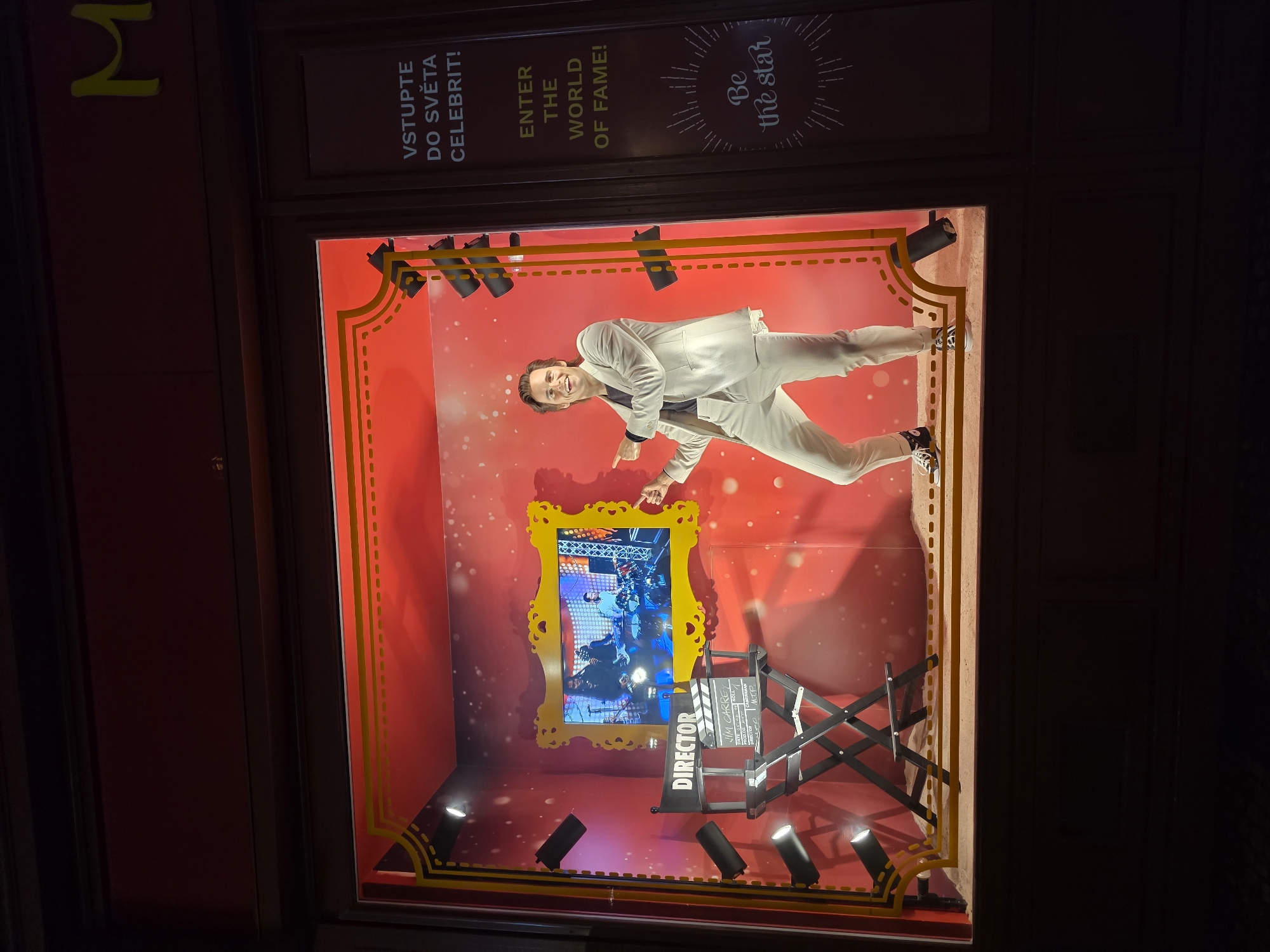 The image shows a store window display featuring a wax figure of Jim Carrey dressed in a white suit. He is striking a pose, pointing to his left. Behind him, a screen displays a scene of musicians performing. A director's chair and a clapperboard are visible in the foreground. The display has a red background with scattered white speckles, resembling stars or confetti, and is framed by yellow decorative elements and theatrical spotlights. To the right of the window, text in Czech reads "VSTUPTE DO SVĚTA CELEBRIT!" and in English, "ENTER THE WORLD OF FAME!" Below this, in script, it says "Be the star." The overall scene evokes a Hollywood or movie premiere theme.