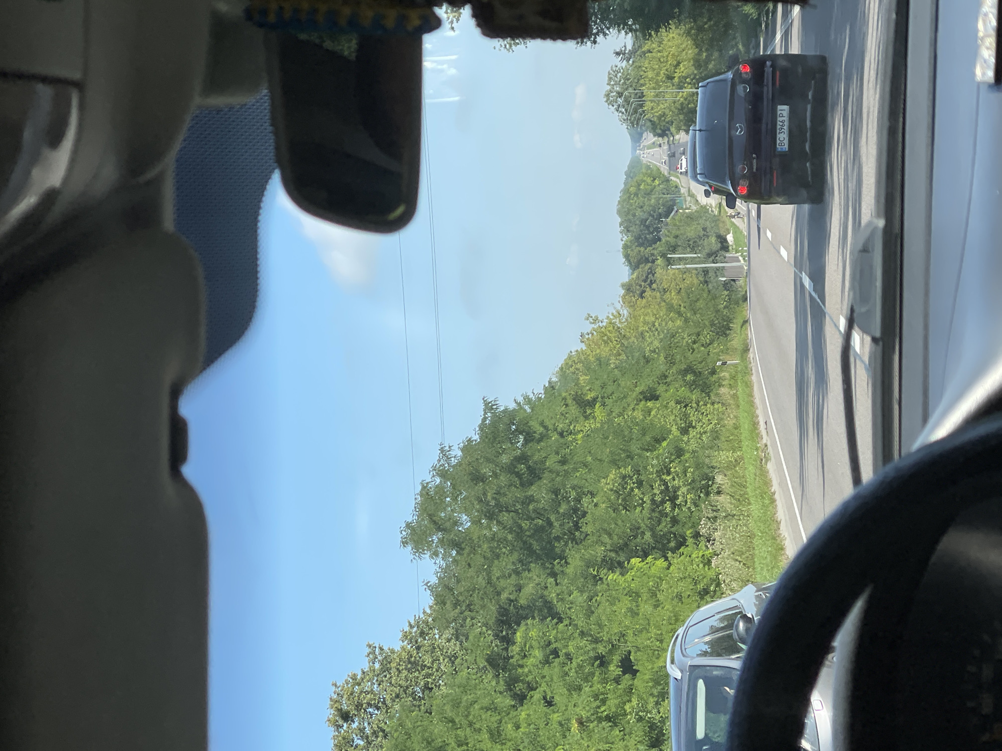 This image, taken from the perspective of someone driving a car, captures a highway scene in Sukhodoly, Ukraine. The sky is a clear blue with a few wispy clouds. Dense green trees line both sides of the road. In the foreground, the steering wheel of the car is visible. On the road ahead, several cars are driving in the same direction. A black Mazda 3, with license plate BC 3966 PI, is in the middle lane, slightly ahead of the viewpoint. Other vehicles are further down the road, receding into the distance. The road is paved and has lane markings. Streetlights are visible along the right side of the road. The overall scene depicts a typical day on a Ukrainian highway.