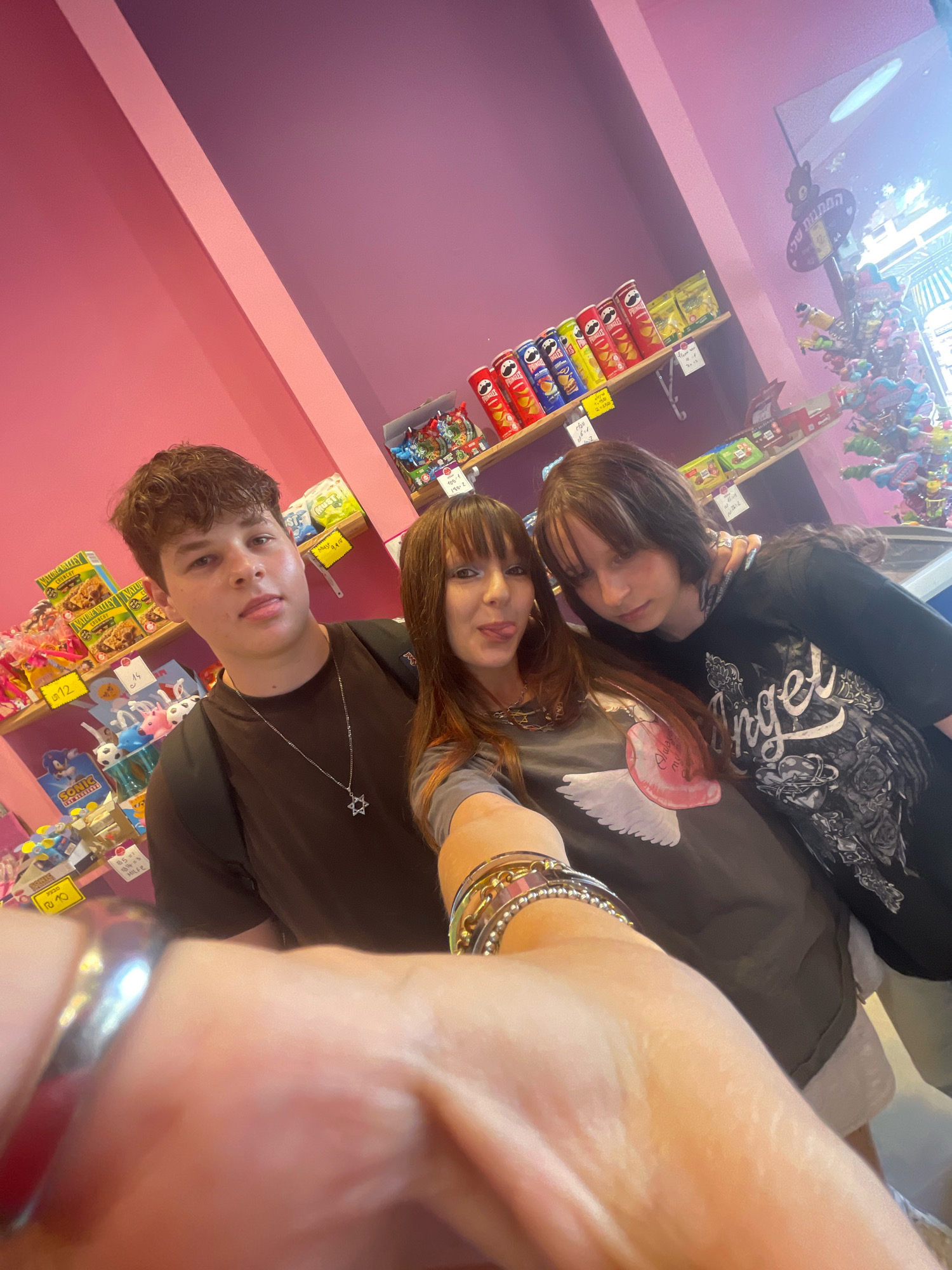 Three individuals are pictured in what appears to be a candy store or a similar retail establishment with shelves stocked with various items, including Pringles cans, bags of snacks, and small toys. The walls are painted in shades of pink and purple, with a diagonal line separating the colors. The store lighting is bright.
The person on the left is a male teenager with short, curly brown hair, wearing a dark t-shirt and a silver Star of David necklace. He is looking at the camera.
The person in the center is a female with long, dark brown hair with bangs, wearing a gray t-shirt with a graphic print of wings and a heart. She is sticking her tongue out towards the camera.
The person on the right is a female with long, dark brown hair with streaks of lighter color, wearing a black t-shirt with a graphic print featuring the word "Angel" and various designs. She is looking down and to her left.
The foreground is dominated by the arm and hand of someone taking the photo, adorned with several bracelets. The shelves behind the people contain a variety of colorful products, suggesting a playful and consumer-oriented environment. One sign on the shelf has Hebrew writing on it, and another sign further up appears to be in Hebrew as well, possibly indicating the location is Raanana, Israel.