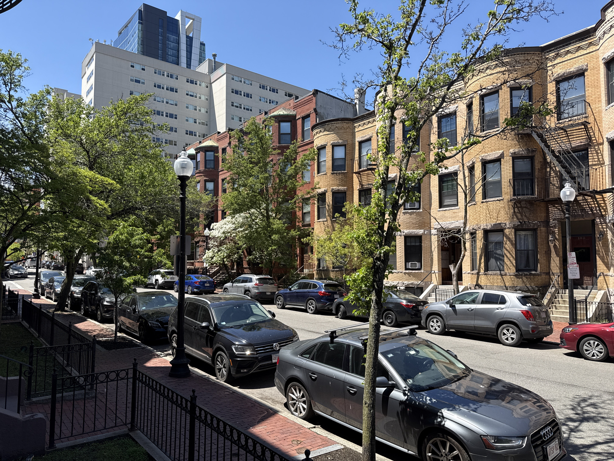 The image shows a residential street in Boston, Massachusetts. The street is lined with parked cars on both sides. The buildings are a mix of brick and brownstone row houses, some with bay windows and fire escapes. A modern high-rise building is visible in the background. Trees are scattered along the street, providing some shade. The weather appears sunny and clear. The street is well-maintained, with brick sidewalks and iron fences.