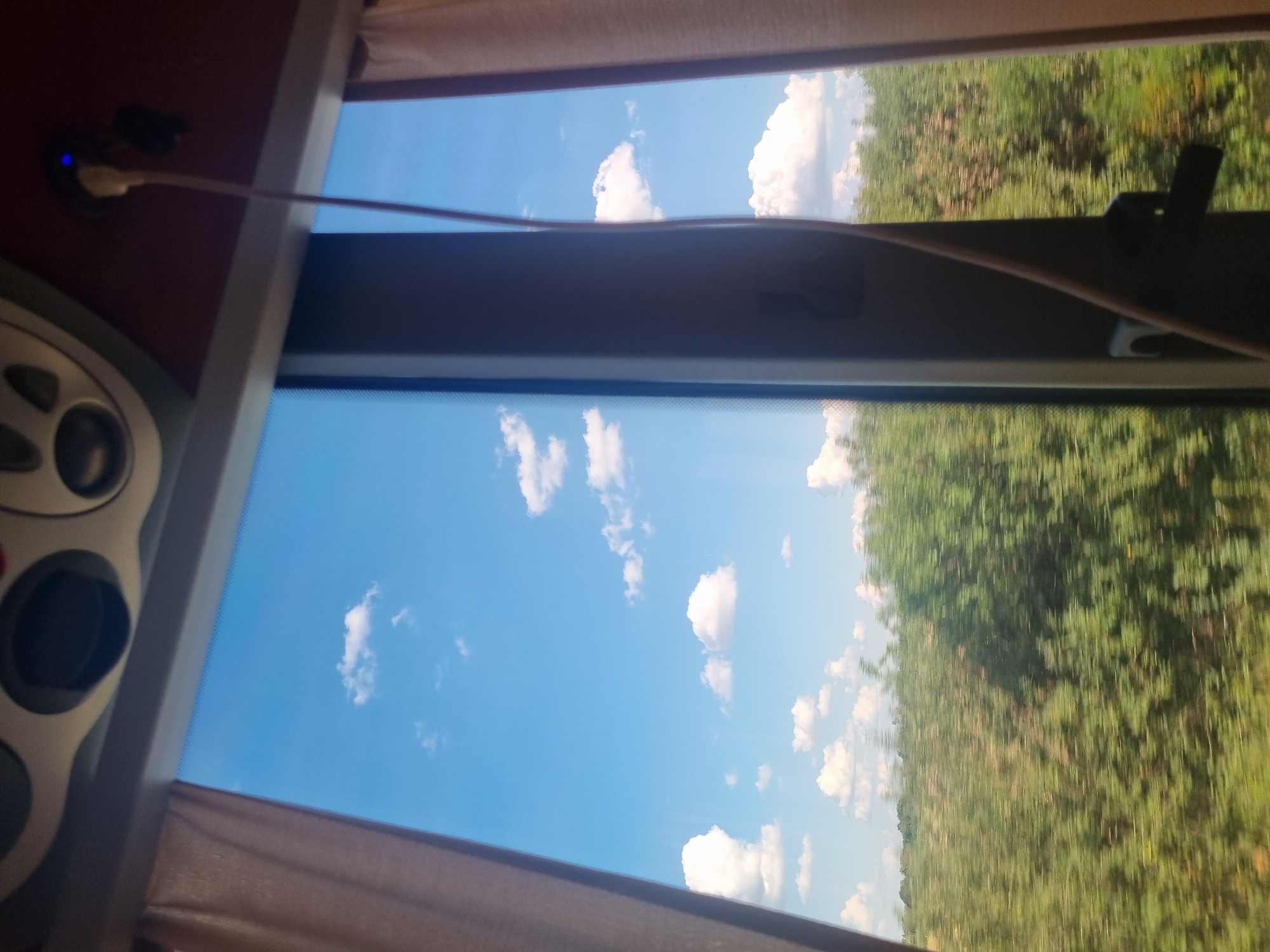 The image shows a view through a window, likely from a moving vehicle. The scene is split into two main sections by a vertical frame. The left side of the window reveals a bright blue sky with scattered white clouds. Below the sky, a blurred green landscape of trees and vegetation is visible, indicating motion. A beige curtain is partially drawn on the far left.
The right side of the window also shows the blue sky and clouds, but it is partially obstructed by a dark, vertical frame. A thin, light-colored cable or wire hangs down from the top right of the frame, attached to a small blue light. Another dark object, possibly a connector or device, is also visible near the top. A portion of a beige curtain is also visible on the far right.
The location of Komoró, Hungary, is not discernible from the visual elements in the image. There are no people or specific urban landmarks present to identify the city or country. The image focuses on the natural scenery outside the window and interior elements of the vehicle.
