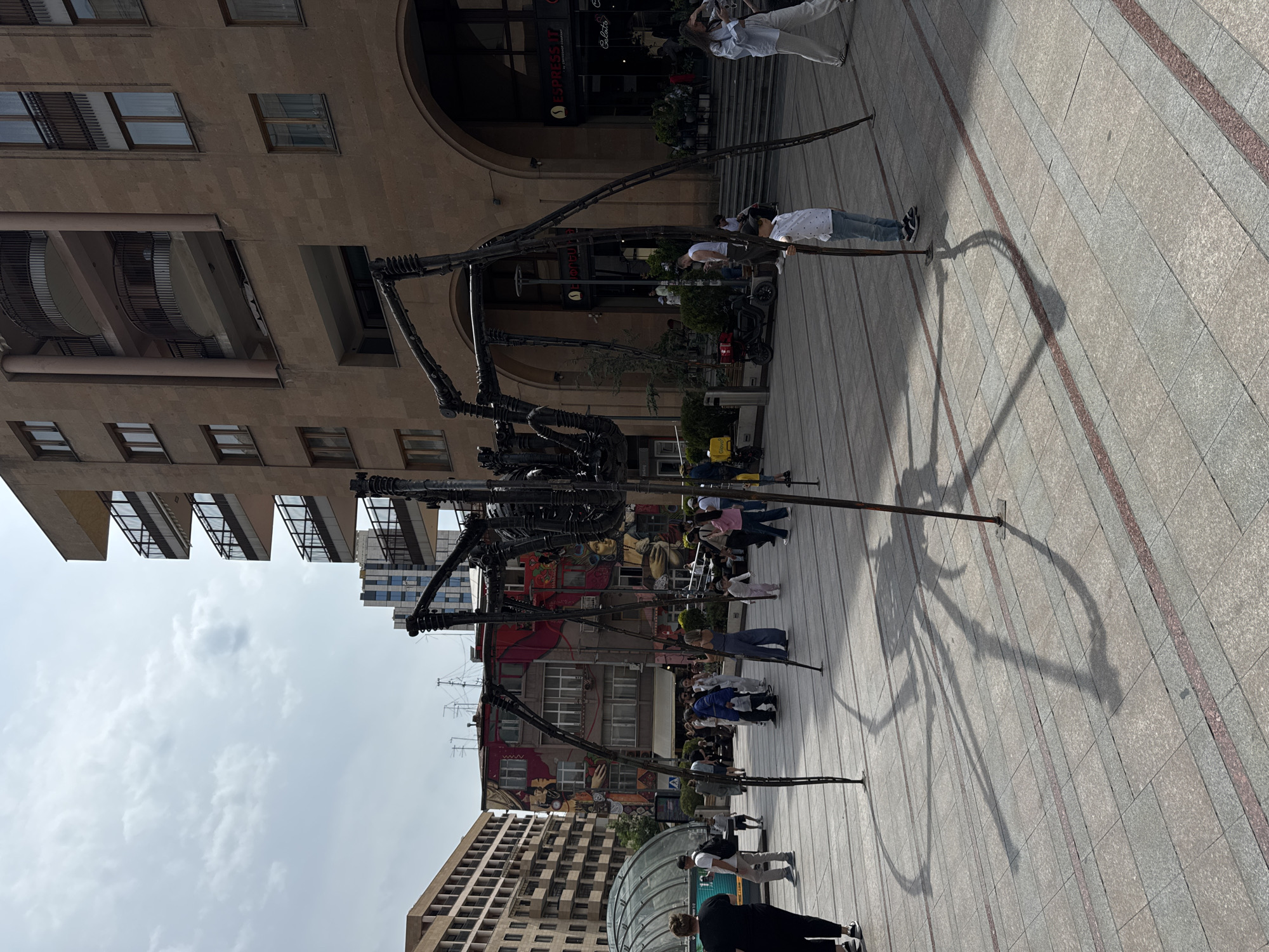 Here is a detailed description of the image:
The image captures a bustling urban scene centered around a large, black, spider-like sculpture standing prominently in a paved square. The sculpture is detailed, with what appear to be articulated legs and a complex body structure. People of various ages are milling around the sculpture, some interacting with it by taking pictures or simply observing. The pavement is made of large, light-colored tiles with linear patterns.
Behind the sculpture, there is a mix of architectural styles. A tall, beige-colored building with multiple floors and archways is visible. Further back, other buildings display various heights and designs, including one with colorful murals painted on its facade.
The sky is overcast, suggesting diffused sunlight. The shadow cast by the sculpture is visible on the pavement, further indicating the presence of light. The image appears to be taken in a public square, with a blend of locals and tourists present. The location is in Yerevan, Armenia.