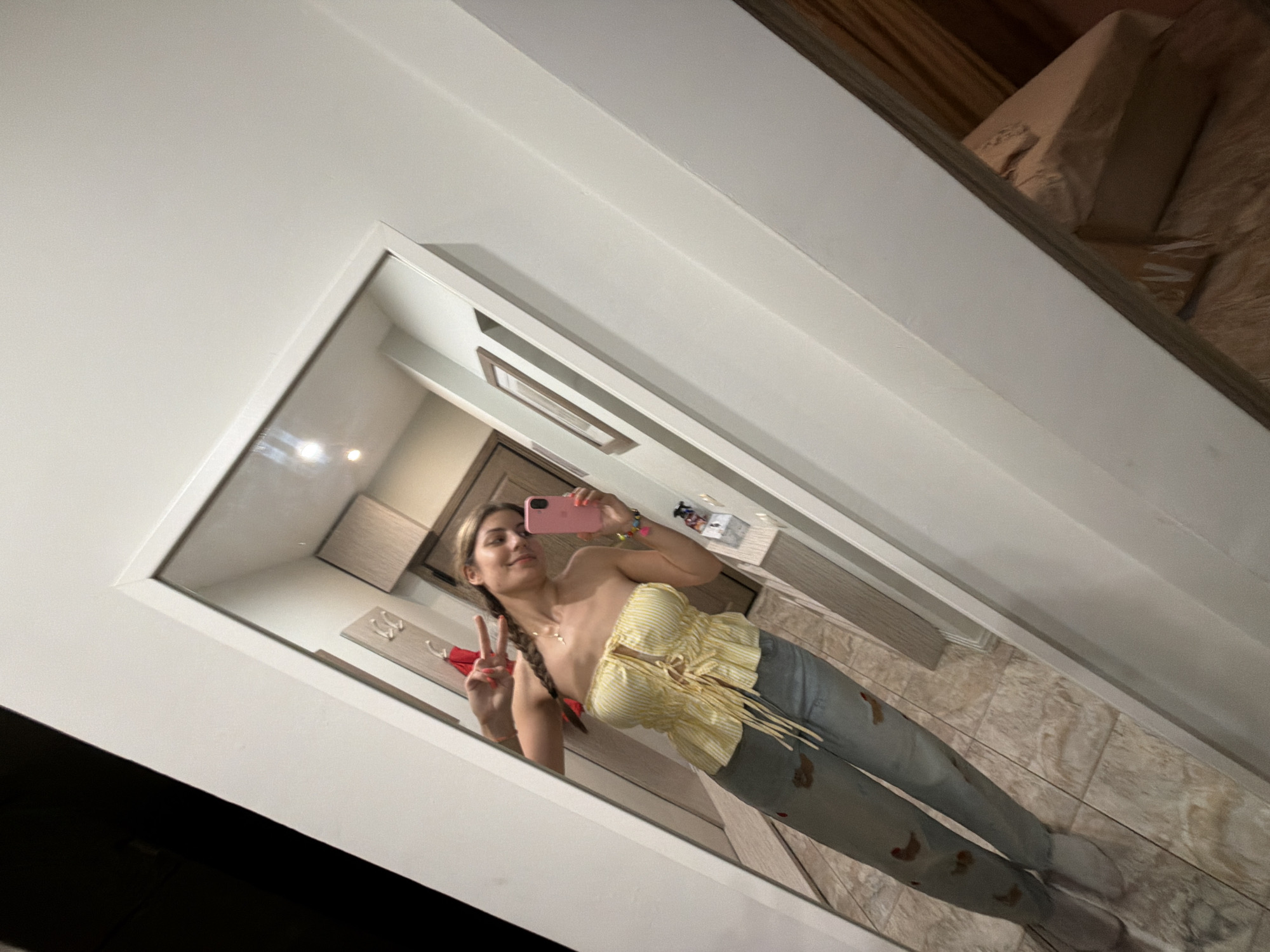 A young woman is captured in a full-length mirror selfie, positioned indoors. She has light brown hair, styled in a braid over her left shoulder, and is wearing a strapless, yellow and white striped top with a cinched, ruffled detail at the waist, and light blue jeans featuring scattered brown patches. She holds a pink smartphone in her right hand to take the photo, while her left hand is raised, making a peace sign. Her expression is a subtle smile, conveying a relaxed or playful mood.
The reflection in the mirror reveals elements of the room's interior. Behind her, a wooden door or cabinet and a small, light-colored shelf with various items, including a red object, are visible. The floor is covered with light-colored, possibly beige or cream, square tiles. In the upper right corner of the mirror's reflection, part of a bed with brown bedding and a headboard can be seen, suggesting a bedroom or hotel room setting. The mirror itself has a clean white frame. The lighting is artificial, illuminating the scene uniformly. No discernible text is visible. The scene is located in Burgas, Bulgaria.