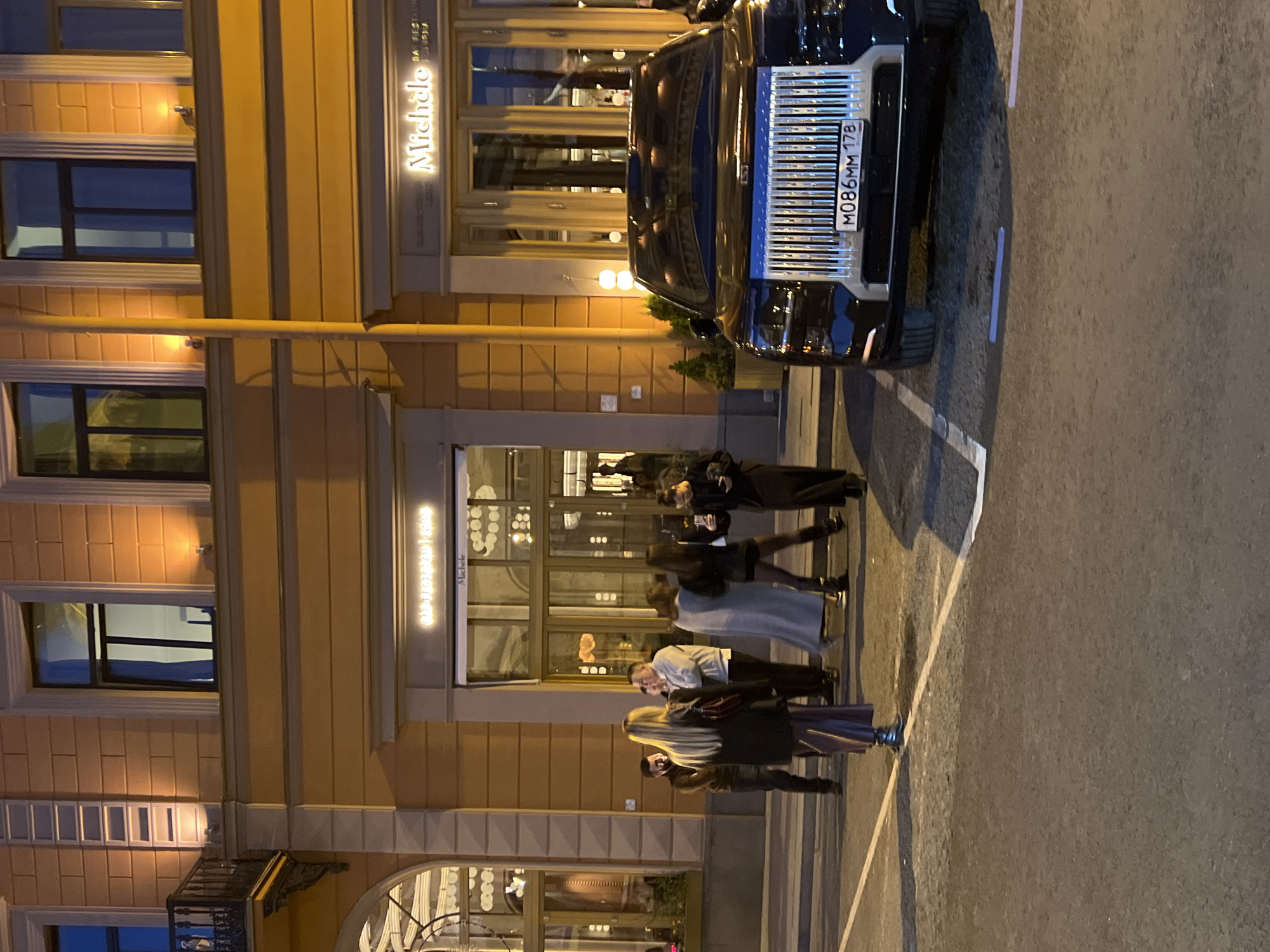 Captured at night, the image shows the exterior of "Michele," a restaurant in Saint Petersburg, Russia. A black luxury car, its license plate "M086MM 178" visible, is parked in front of the establishment. A small group of people is gathered near the restaurant entrance. The architecture features large windows, decorative molding, and illuminated signage. The scene conveys a sense of upscale dining and nightlife.