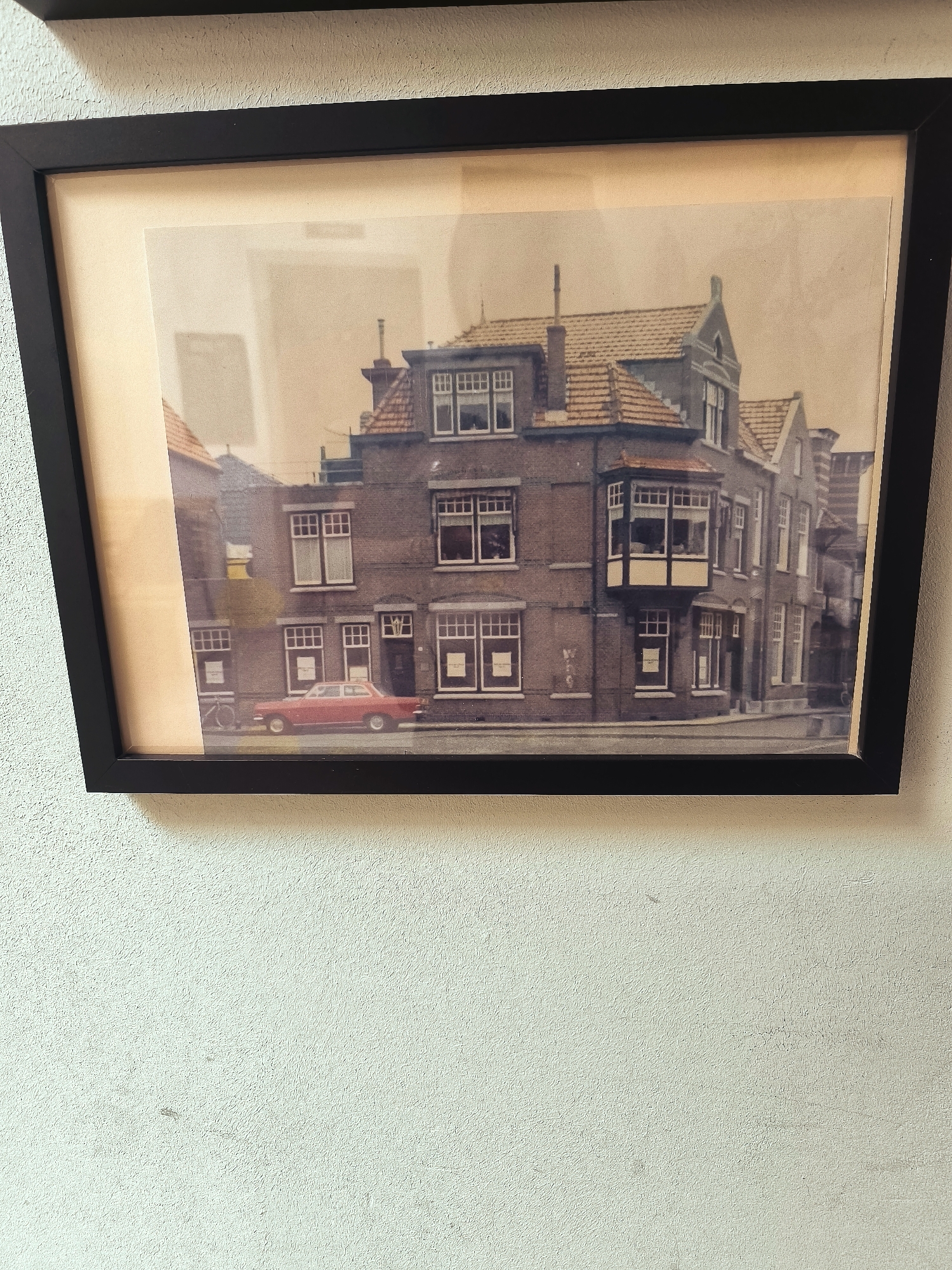The framed photograph shows a vintage image of a street in Hoorn, Netherlands. The primary subject is a red car parked on the street in front of a brick building. The building appears to be a two or three-story structure with multiple windows and a tiled roof. There is a bay window on the second floor. A bicycle is leaning against the building on the left side of the image. The street is paved, and there are some faint markings or graffiti on the building. The overall scene suggests a quiet street in a European city. The photograph itself is framed in black and has a light-colored mat. The wall on which the picture is hanging has a textured surface.