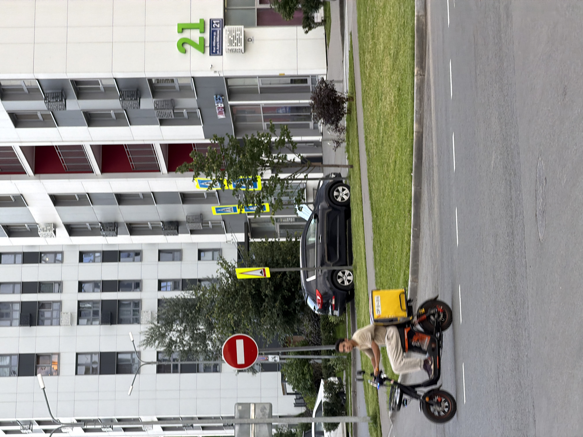 A person on a scooter is driving on a street in front of a modern building. The scooter is black and has a yellow box on the back. The person is wearing a light brown t-shirt and light-colored pants. The building is tall and has a white facade with gray and red accents. There are windows on the building and an air conditioner unit. A red and white stop sign is visible on the left side of the image. A black SUV is parked on the side of the road. The street is paved and there is a green grassy area next to it. The sky is overcast.