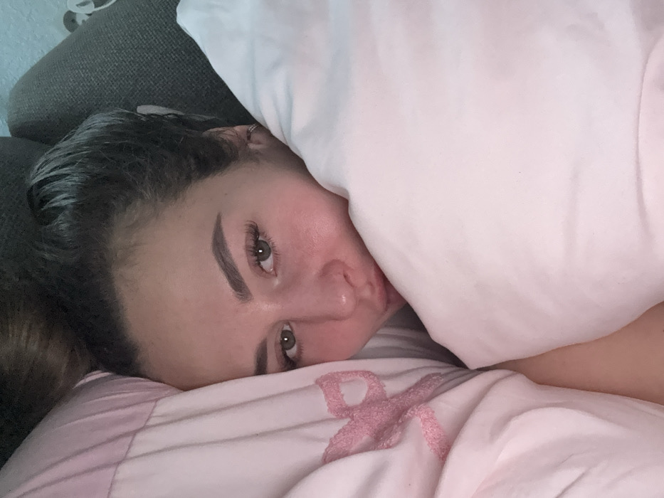 A close-up image features a female individual indoors, partially enveloped by light pink bedding. Her head is positioned against a pillow, with a dark, textured surface visible behind her upper right side. Her dark hair appears pulled back. The subject exhibits fair skin, defined eyebrows, and dark eyes with eyelash extensions. Her lips are subtly pursed. A small hoop earring is present in her visible right ear. To her left, a section of pink bedding displays an embossed, ribbon-like pattern. A plain light pink comforter covers her lower face and torso on the right. The environment is consistent with a bedroom. No visual cues within the image delineate a specific location in Kyiv, Ukraine.