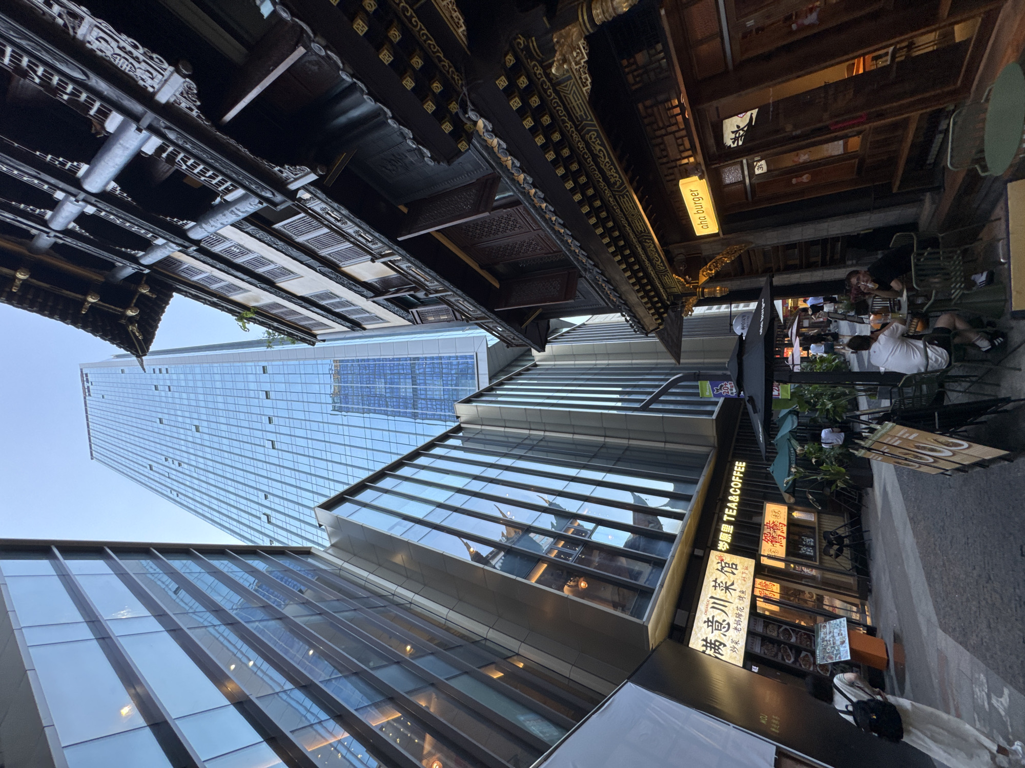 This is a low-angle shot of a street scene in Yuzhong District, China, featuring a juxtaposition of modern and traditional architecture under a clear sky. On the left, a contemporary skyscraper with a glass facade dominates the upper frame, reflecting the sky and surrounding buildings. In the mid-ground, a lower, angular modern building also with a glass exterior is present. To the right, an older, multi-story building with traditional Chinese architectural elements, including dark wood detailing, ornate carvings, and tiled roofs, stands in stark contrast to the modern structures. The street level is lined with businesses, including a restaurant with Chinese characters on its signage and a "aha burger" establishment with a glowing yellow sign. Several people are visible in the scene: a woman in a light-colored dress and backpack walks away from the camera on the left, and two men are seated at a table outside the burger restaurant on the right. Other individuals are seen walking in the background. The ground is paved, and outdoor seating with umbrellas suggests a commercial area.