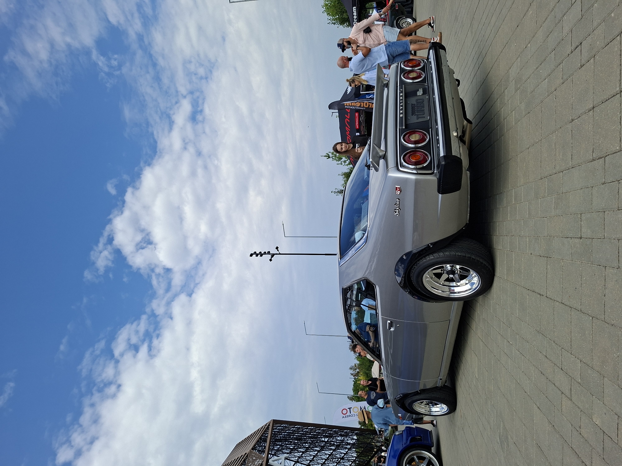An outdoor car show is taking place on a paved area in Stăuceni, Moldova, under a partially cloudy blue sky. The main subject is a silver vintage Nissan Skyline GT-R, viewed from the rear and slightly to the left. The car has a prominent rear spoiler and distinctive round taillights. Its license plate reads "SKYLINE GT TURBO 2000GT".
In the foreground, the paved ground is made of rectangular grey stones. To the left of the Skyline, a portion of a blue sports car is visible. Several people are scattered around the scene, some observing the cars and others interacting.
To the left of the frame, a modern, angular building with a latticed exterior is present. Several tents and flags are set up in the background, suggesting an event area. People are dressed casually in warm-weather clothing. The overall atmosphere appears to be a public gathering focused on automotive enthusiasts.