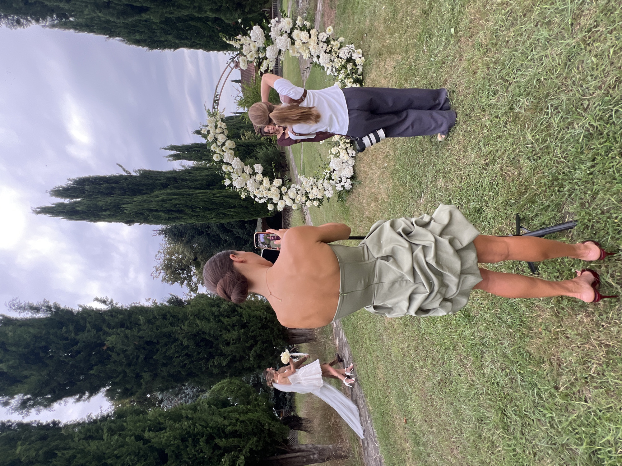 In the city of Napareuli, Georgia, a bride in a short white dress and veil walks across a grassy path flanked by tall cypress trees. A man in a dark suit walks beside her. In the foreground, a woman in a green strapless dress is taking a photo of the bride with her phone. Further back, another woman in a white shirt and dark pants stands with a camera around her neck, facing a circular floral arch adorned with white flowers. The sky is overcast.